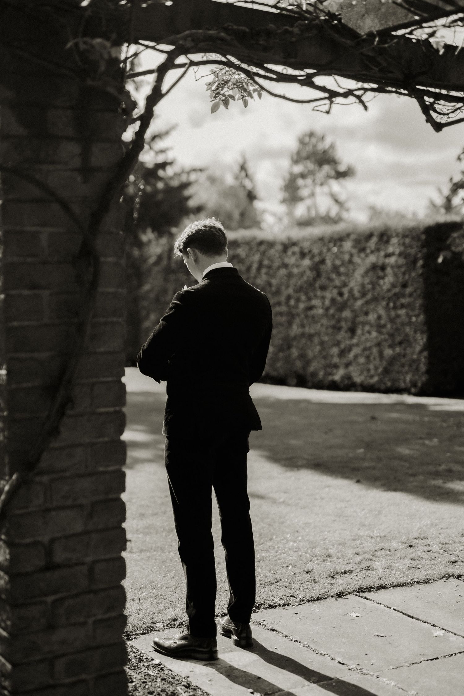 Groom in black suit standing alone under pergola in garden, black and white wedding photography.