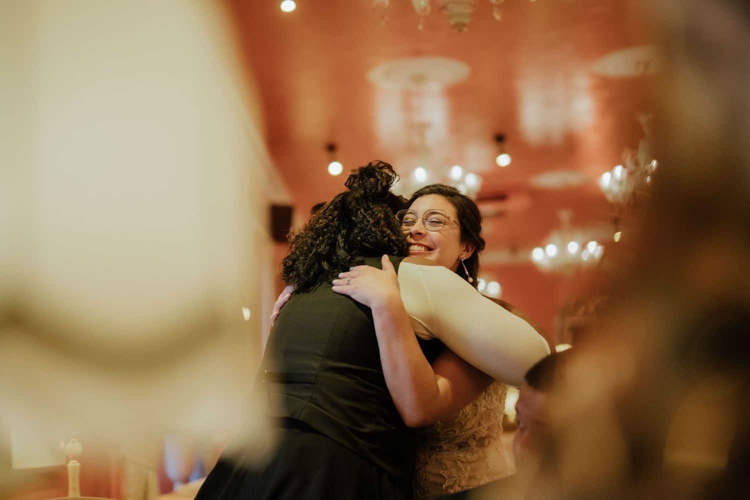 Bride in glasses shares warm embrace with guest at wedding reception with string lights.