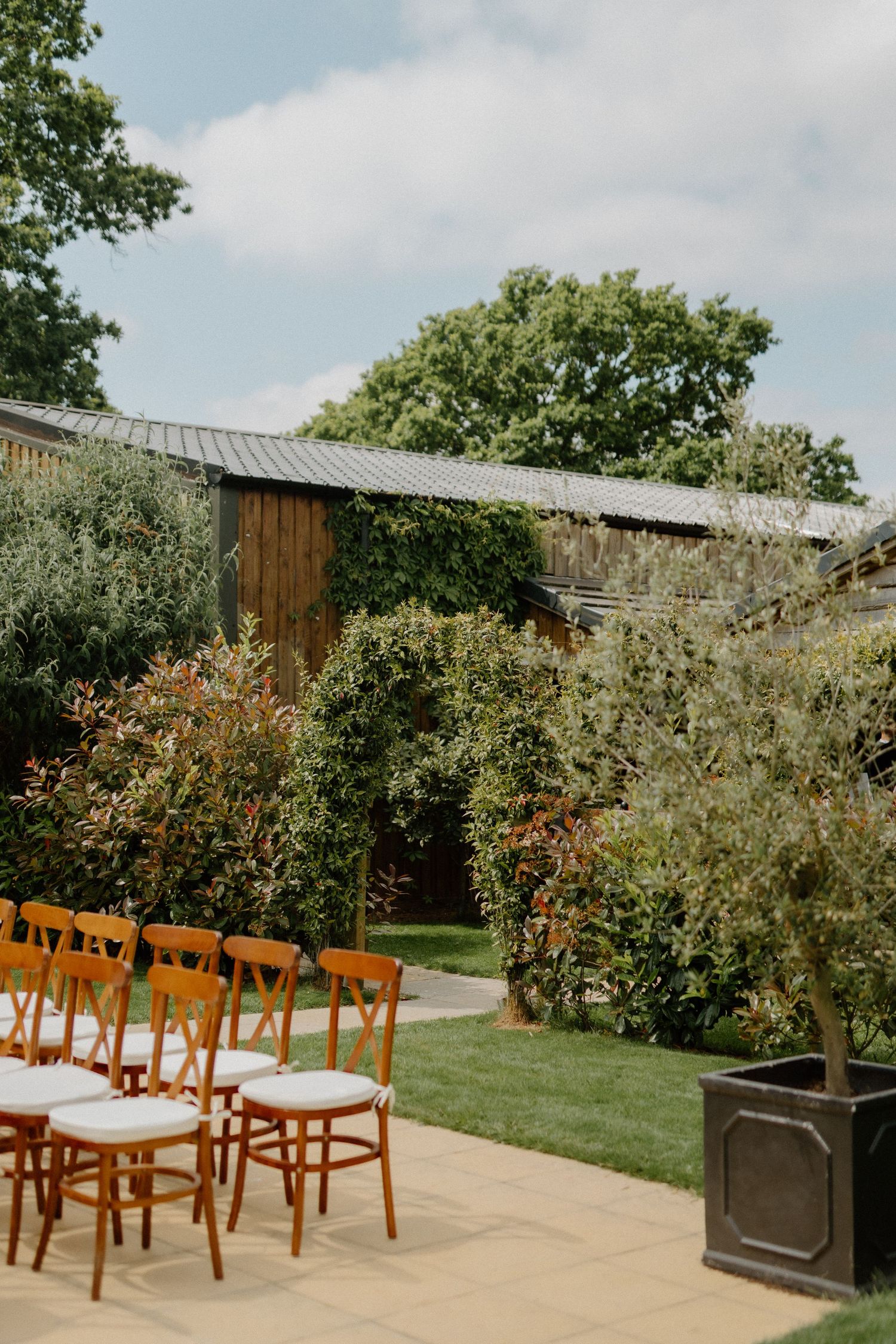 Outdoor wedding ceremony venue with wooden chairs on a sunny day surrounded by lush green gardens.