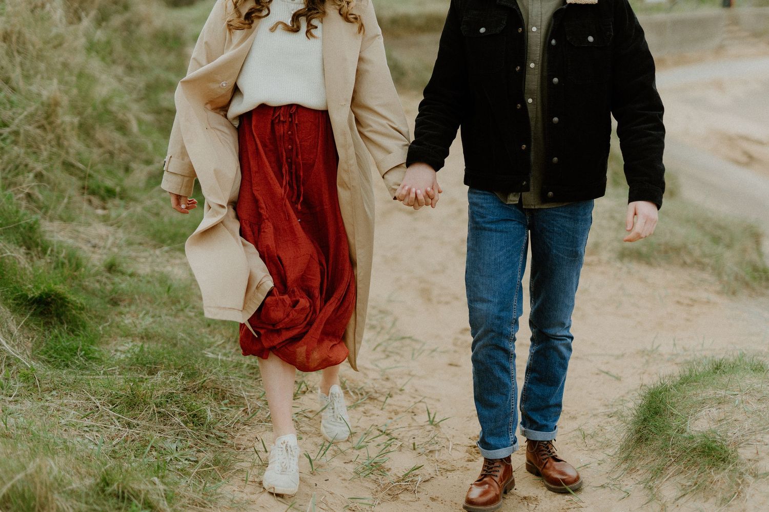 Couple holding hands walking on sandy coastal path, woman in red skirt and beige coat with man.