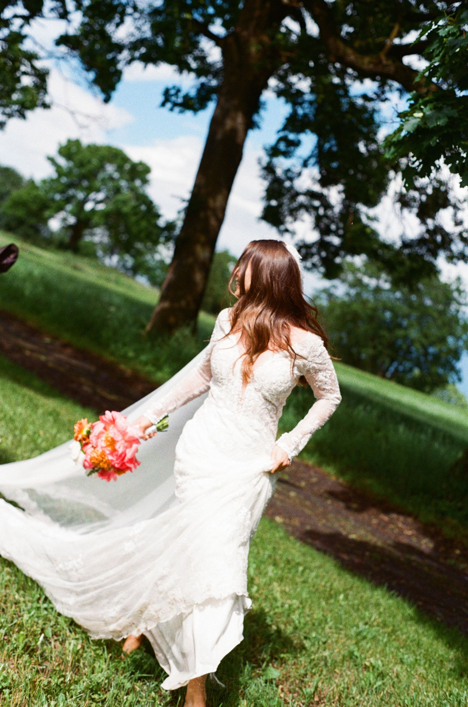 Bride in lace gown twirling with a vibrant bouquet on a green hillside under trees.