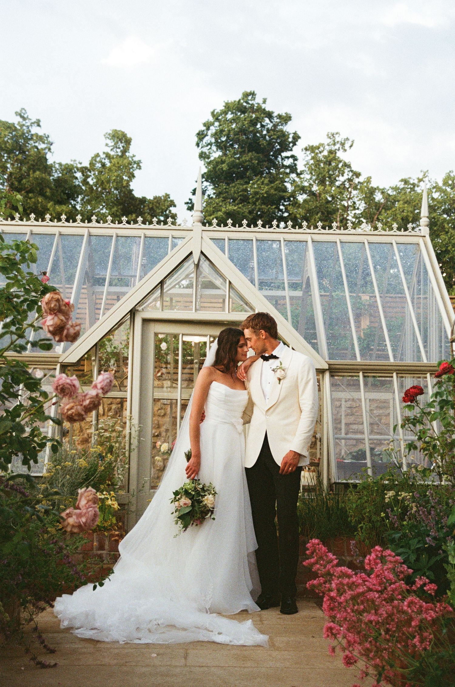 Bride and groom share a tender moment in front of a charming glass greenhouse surrounded by roses.