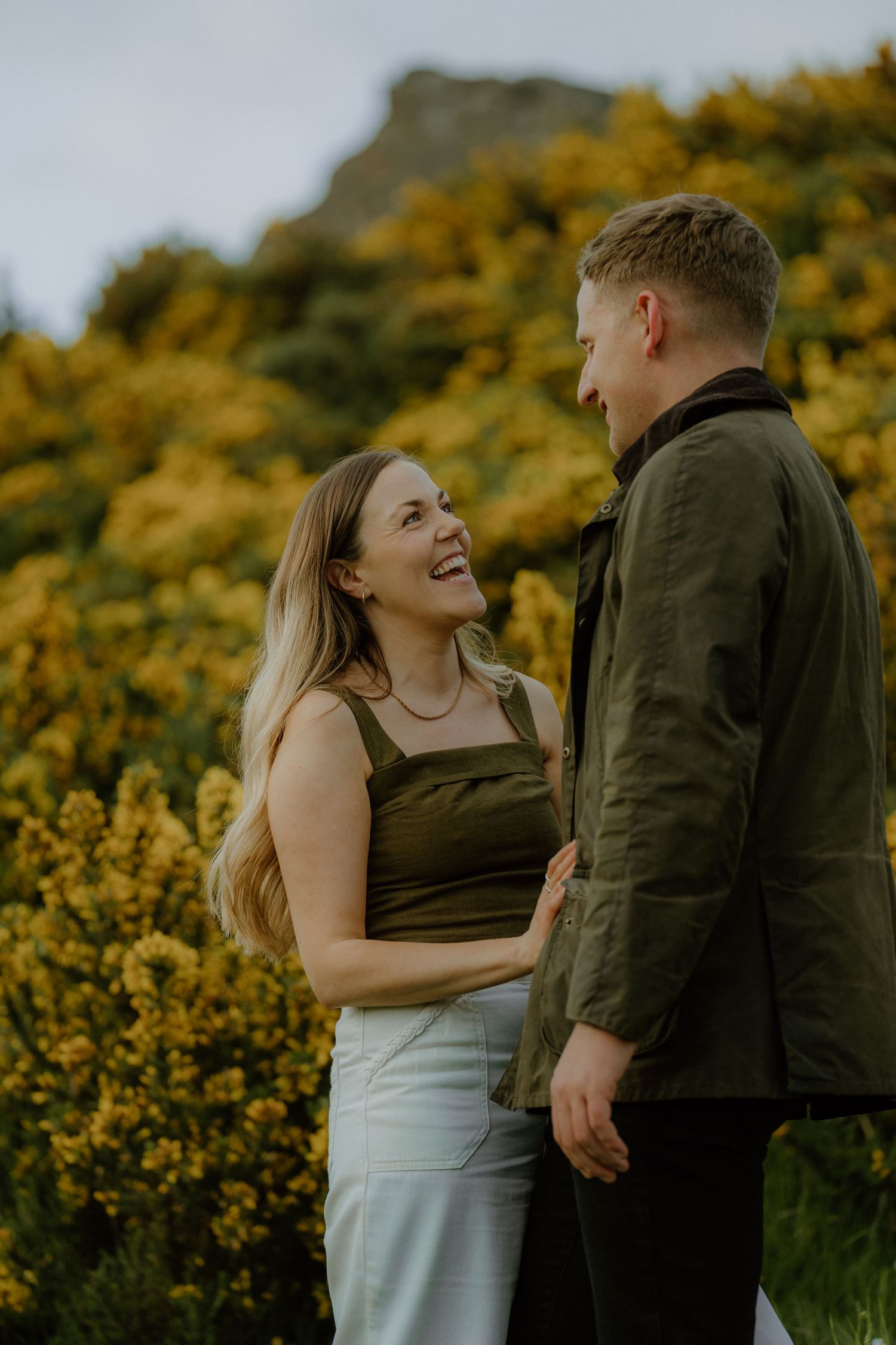 Laughing couple standing together among yellow gorse bushes with rocky hillside backdrop during outdoor engagement shoot.