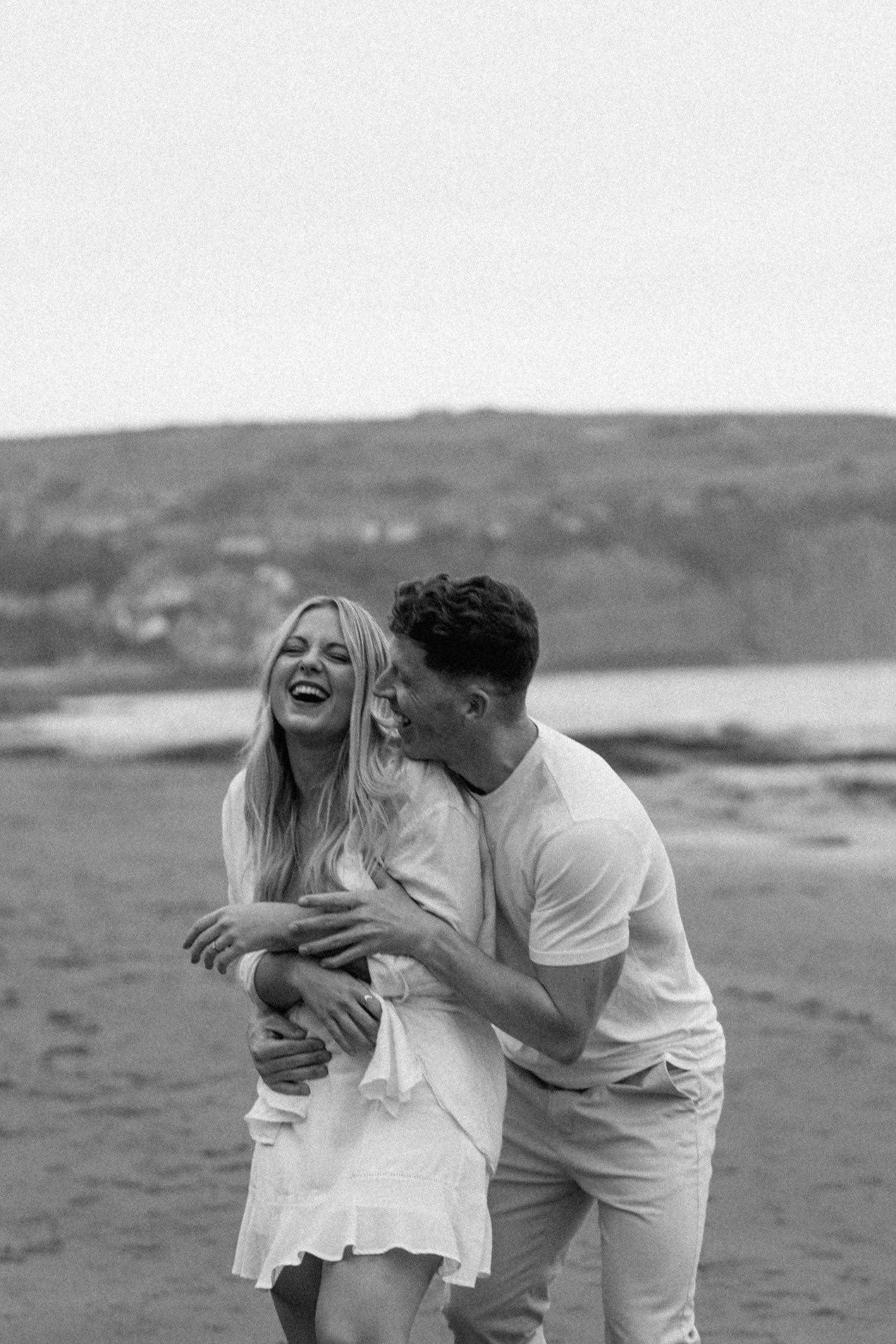 Black and white photo of a man kissing a laughing woman on a sandy beach.