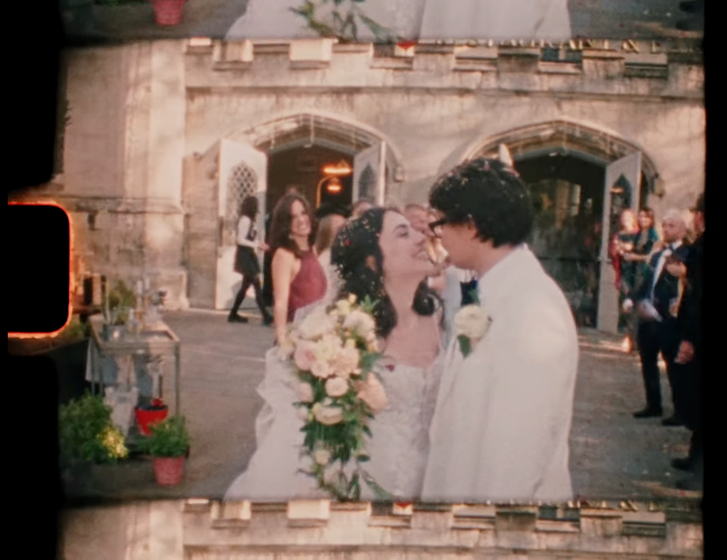 Newlywed couple sharing a kiss outside a stone church, bride holding a bouquet, guests celebrating in the background.