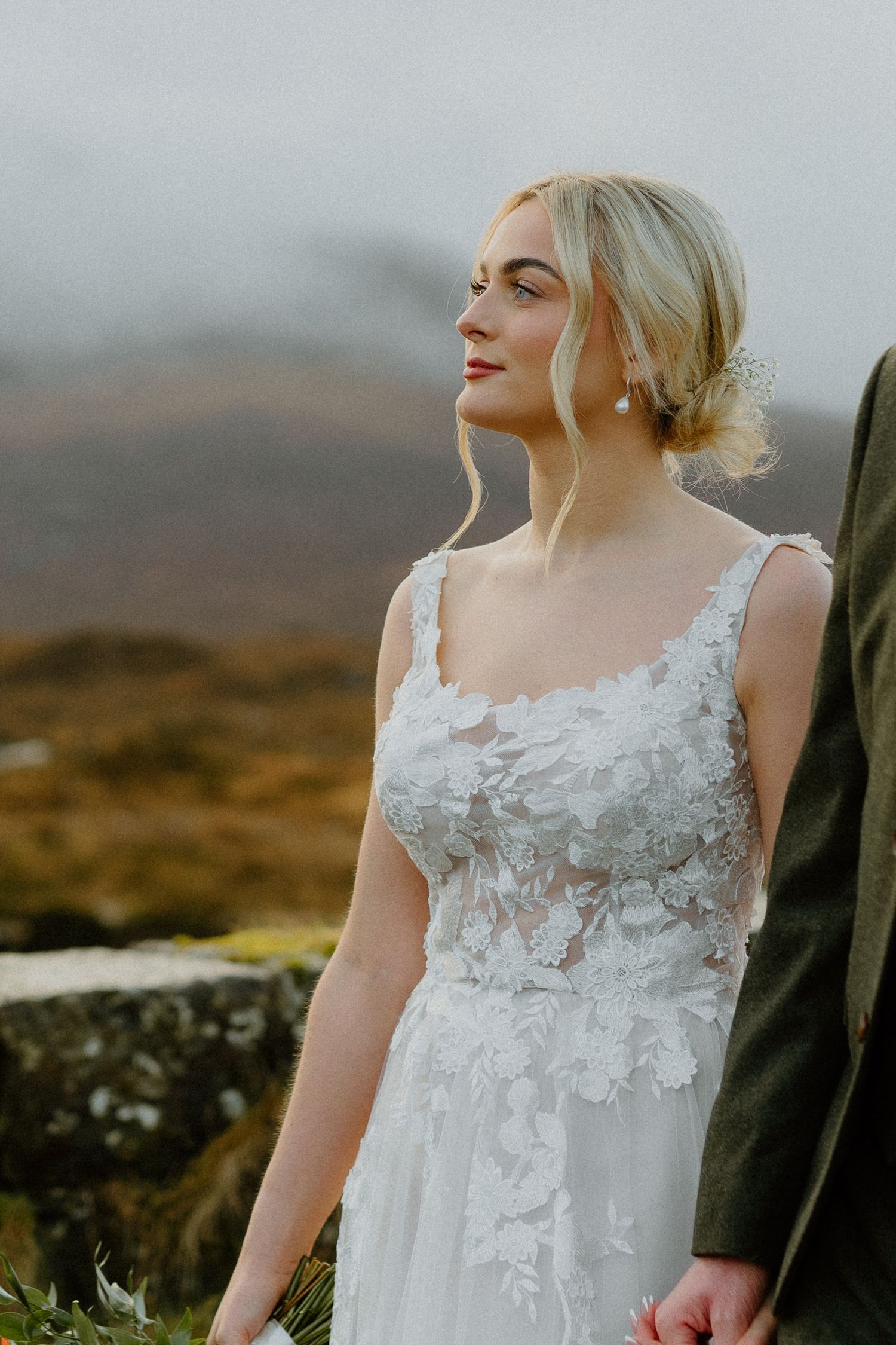 Close-up of radiant blonde bride in floral applique wedding gown gazing serenely against misty Scottish Highland mountains.