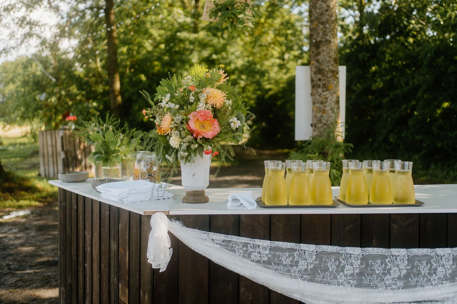 Outdoor wedding welcome table with lace cloth, floral arrangement, and glasses of lemonade.