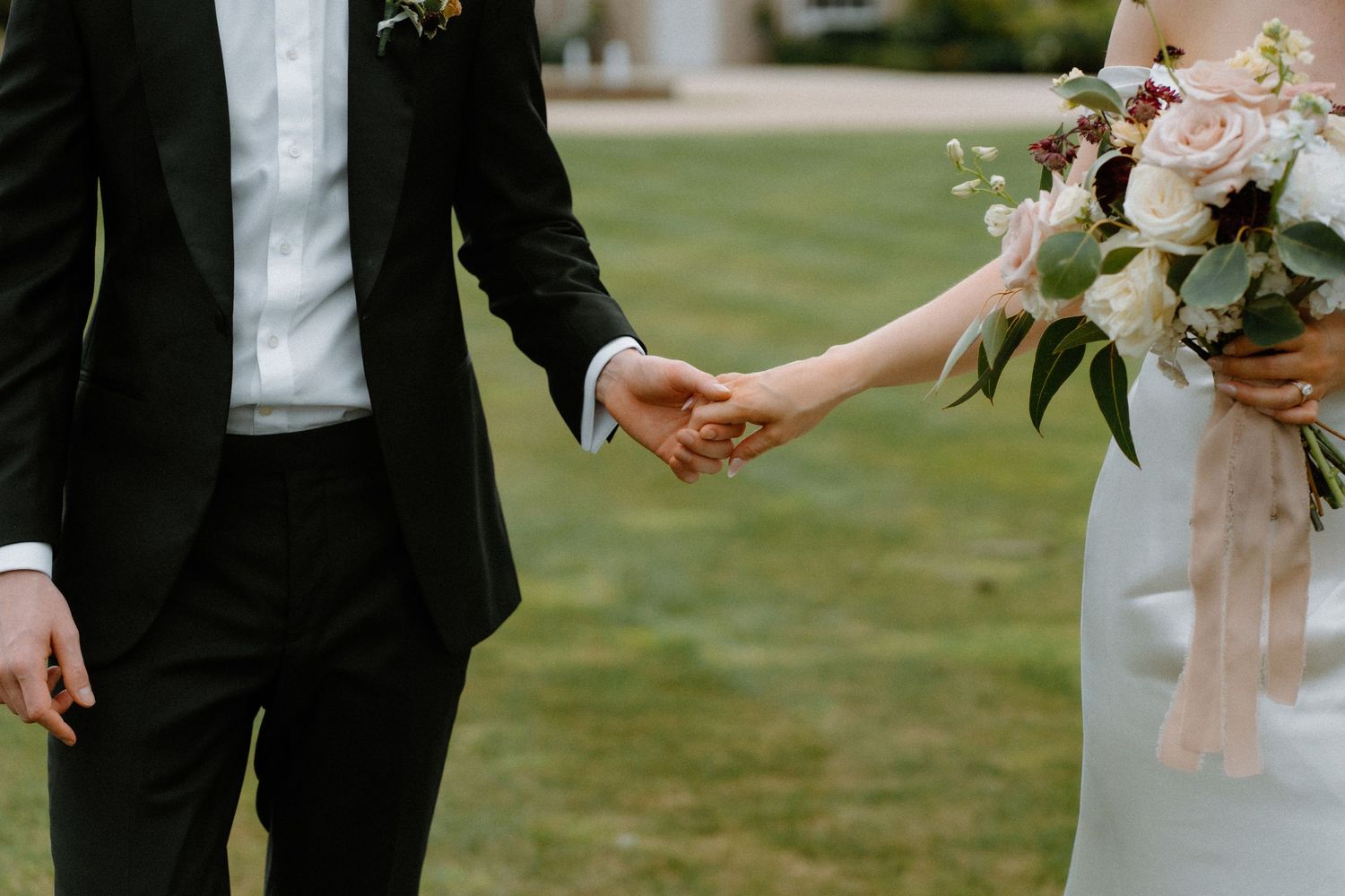 Bride and groom holding hands outdoors, bride carrying a romantic bouquet of blush and burgundy flowers.