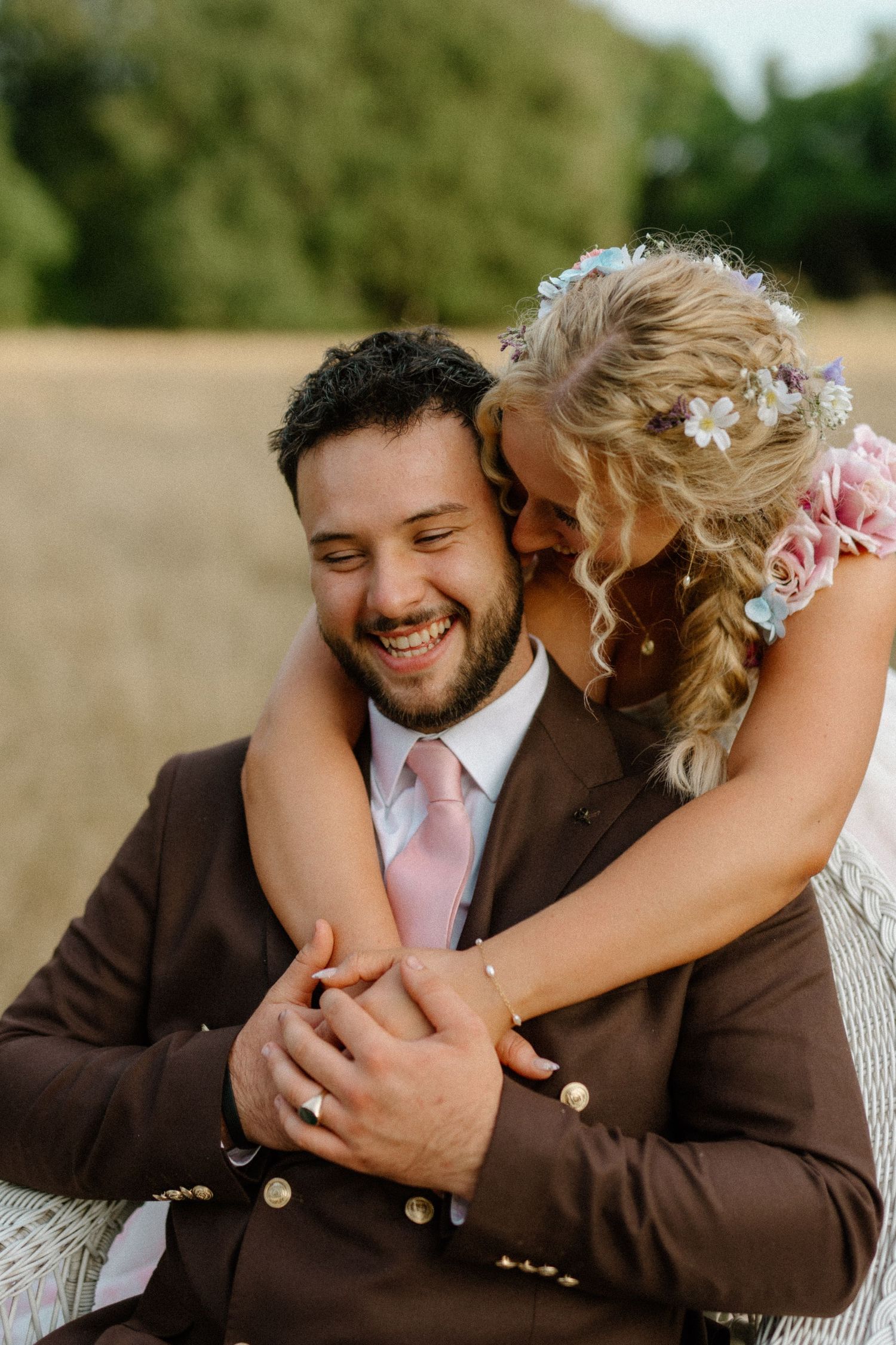 Groom in brown suit laughs joyfully as bride in floral gown embraces him from behind in golden field.