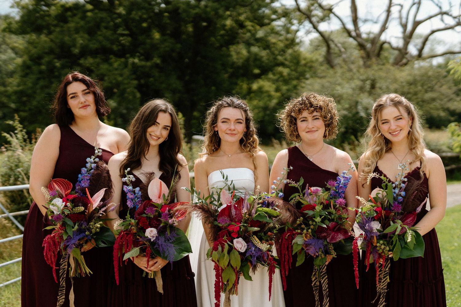 Bride with five bridesmaids in burgundy gowns holding vibrant jewel-toned floral bouquets outdoors.