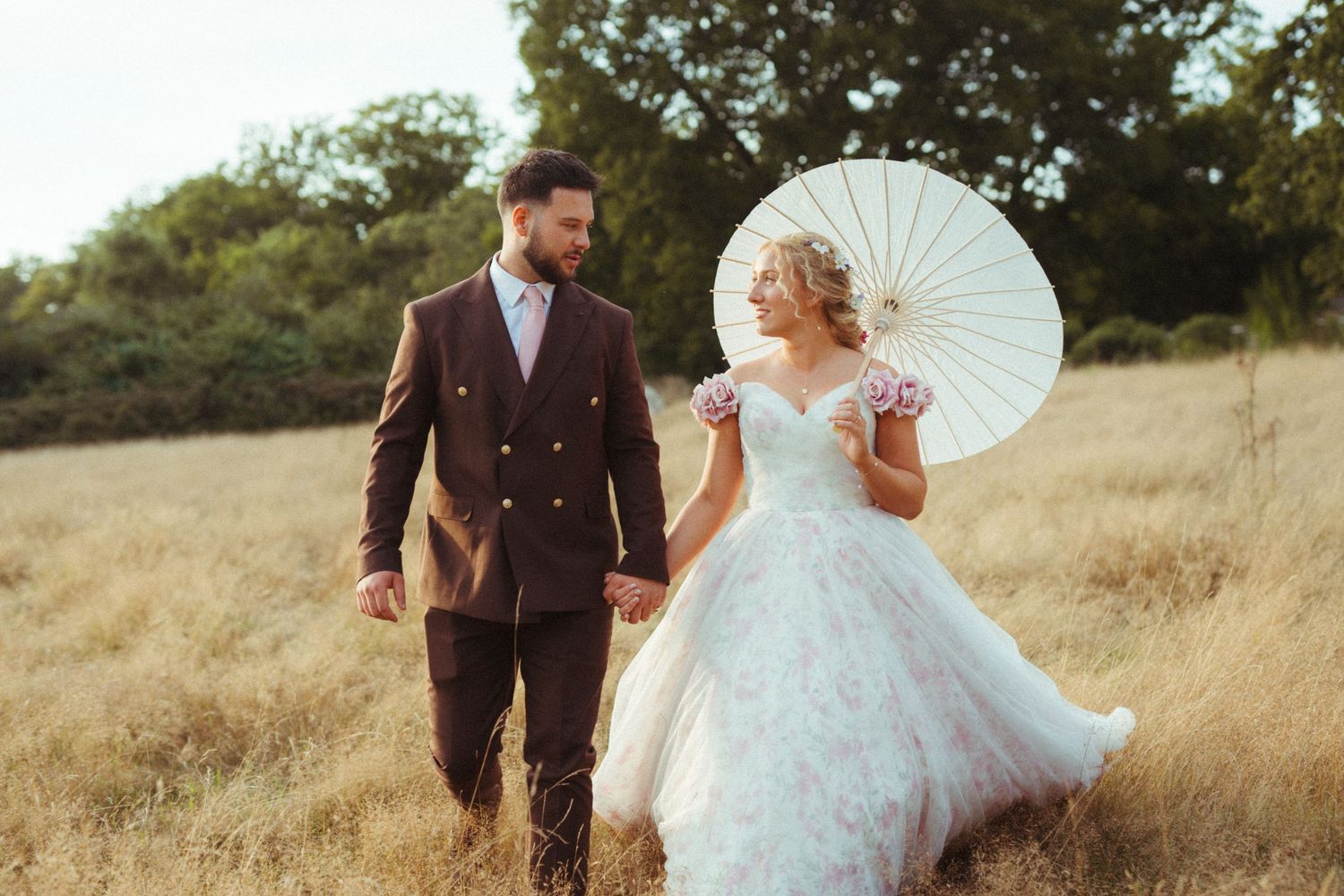 Bride in ball gown with white parasol and groom in burgundy suit walk through golden field at sunset.