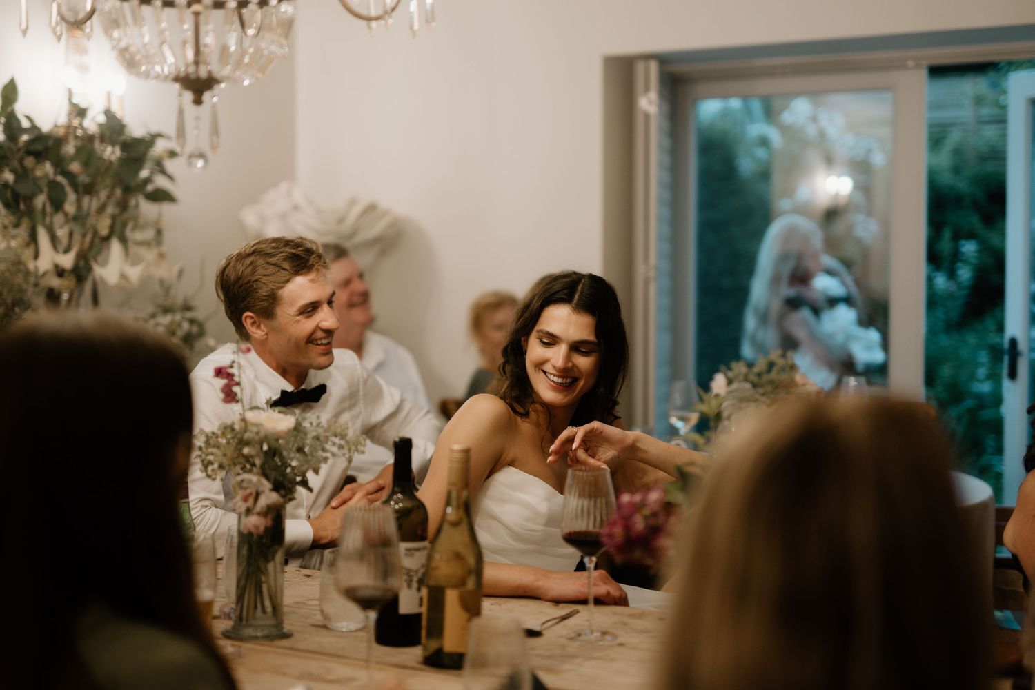 Bride and groom laughing together at wedding reception dinner table with guests.