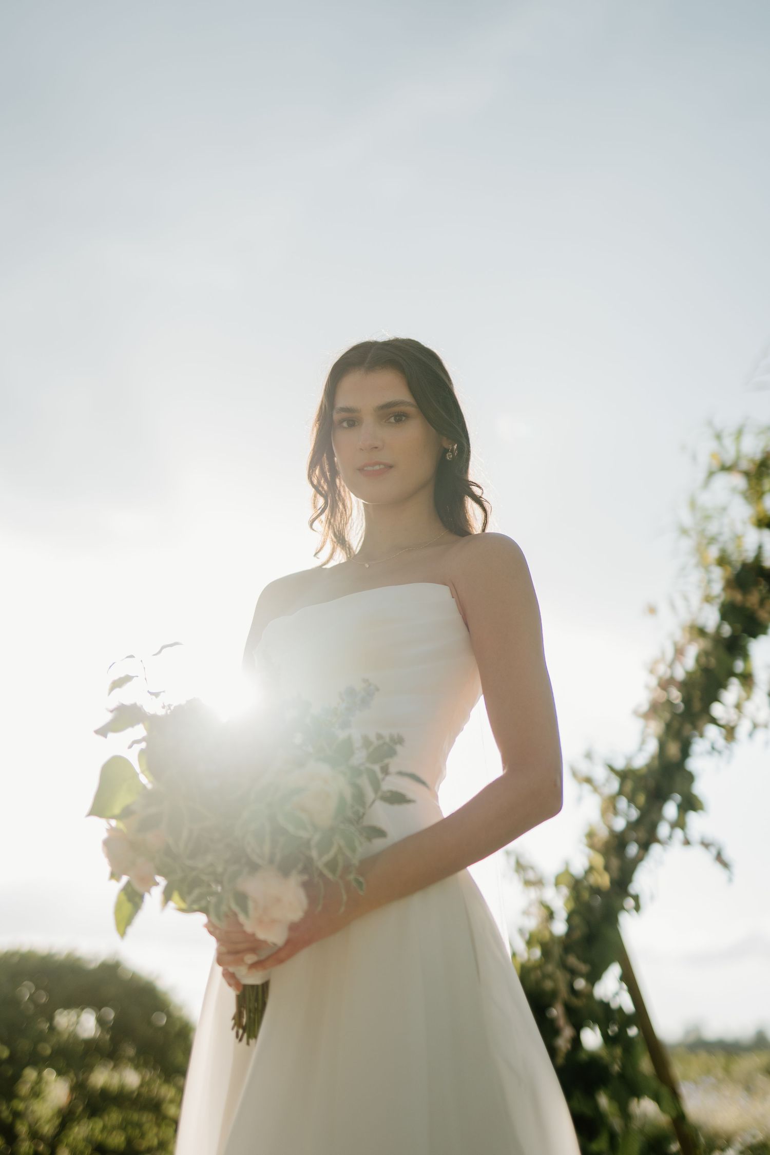 Bride in strapless white gown holds floral bouquet with sunlight glowing behind her against a cloudy sky.