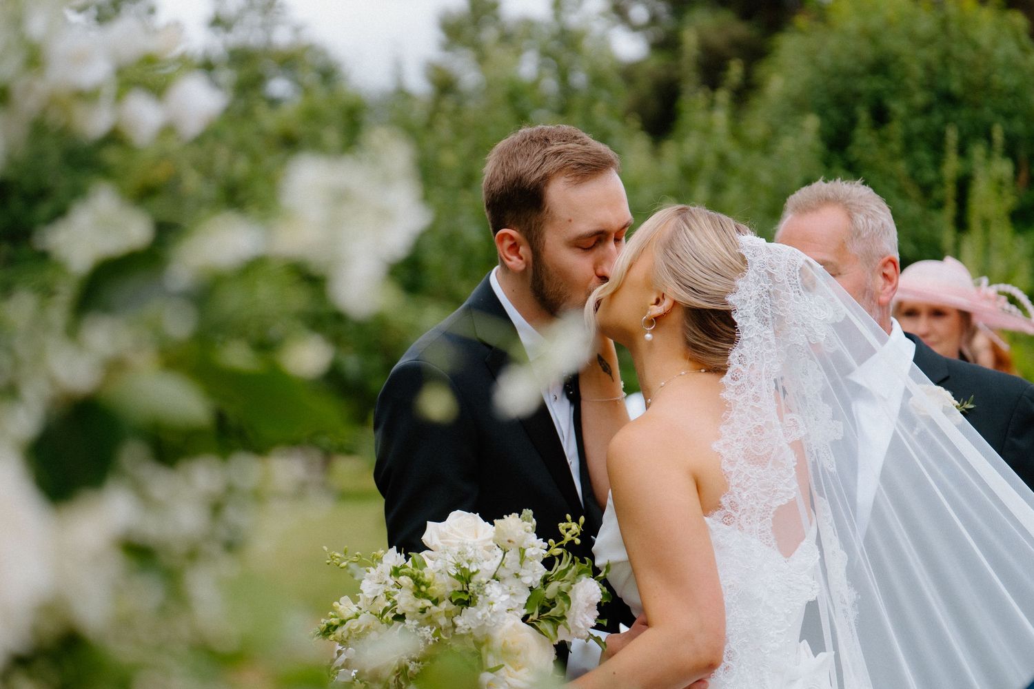 Newlyweds share first kiss surrounded by lush white floral arrangements at outdoor garden wedding.