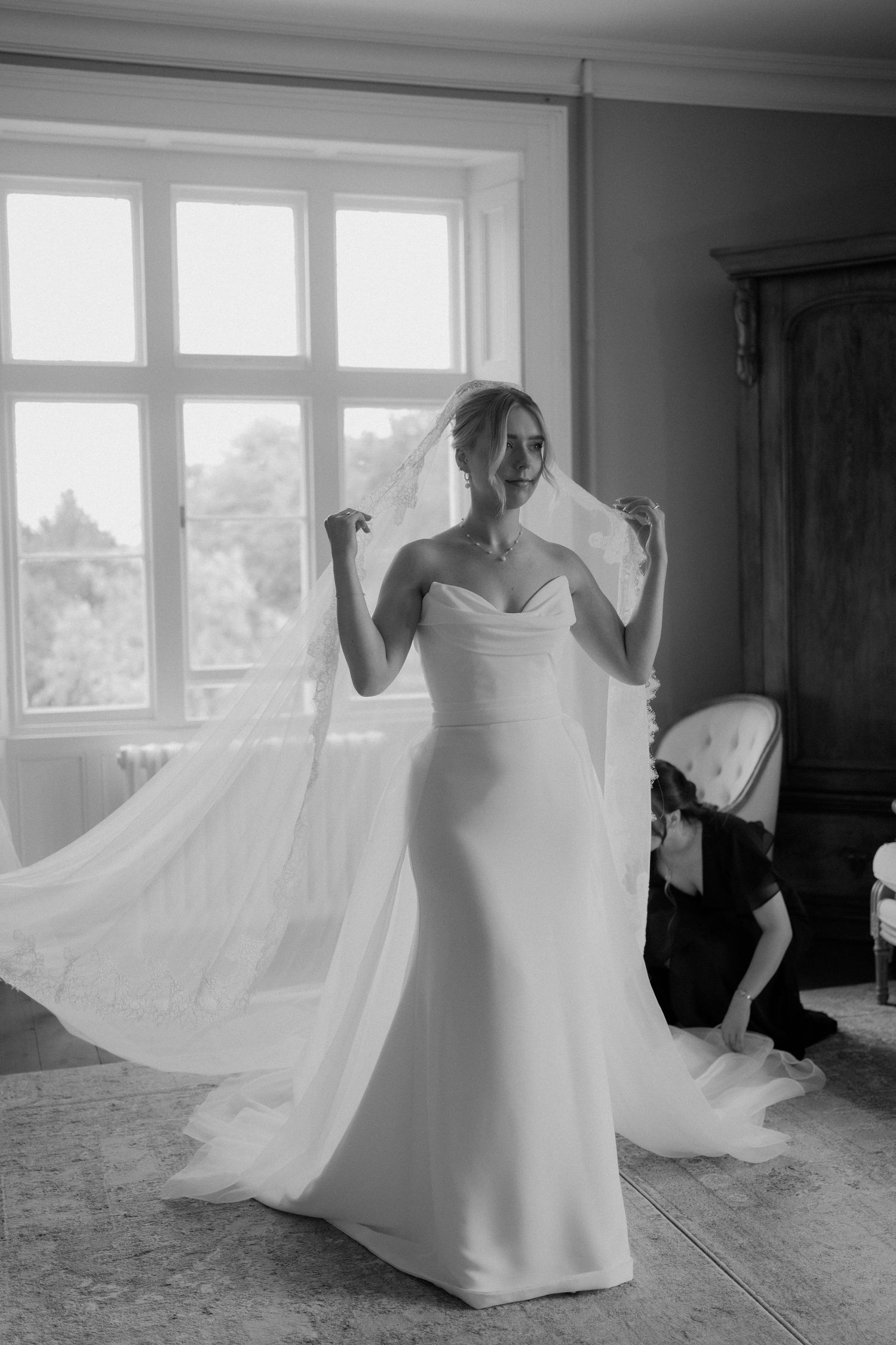 Black and white photo of bride in strapless gown holding veil near large window while being helped.