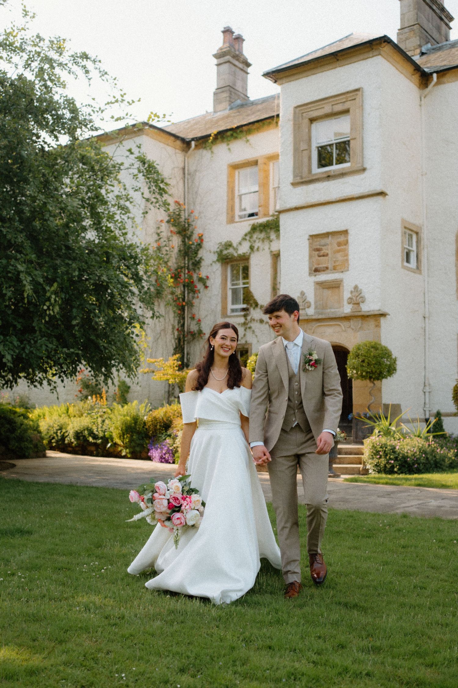 Happy couple walking hand-in-hand across lawn in front of grand white country house wedding venue.