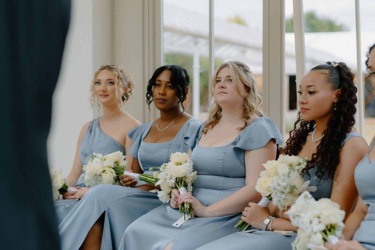 Four bridesmaids in dusty blue dresses holding white bouquets at a wedding ceremony.