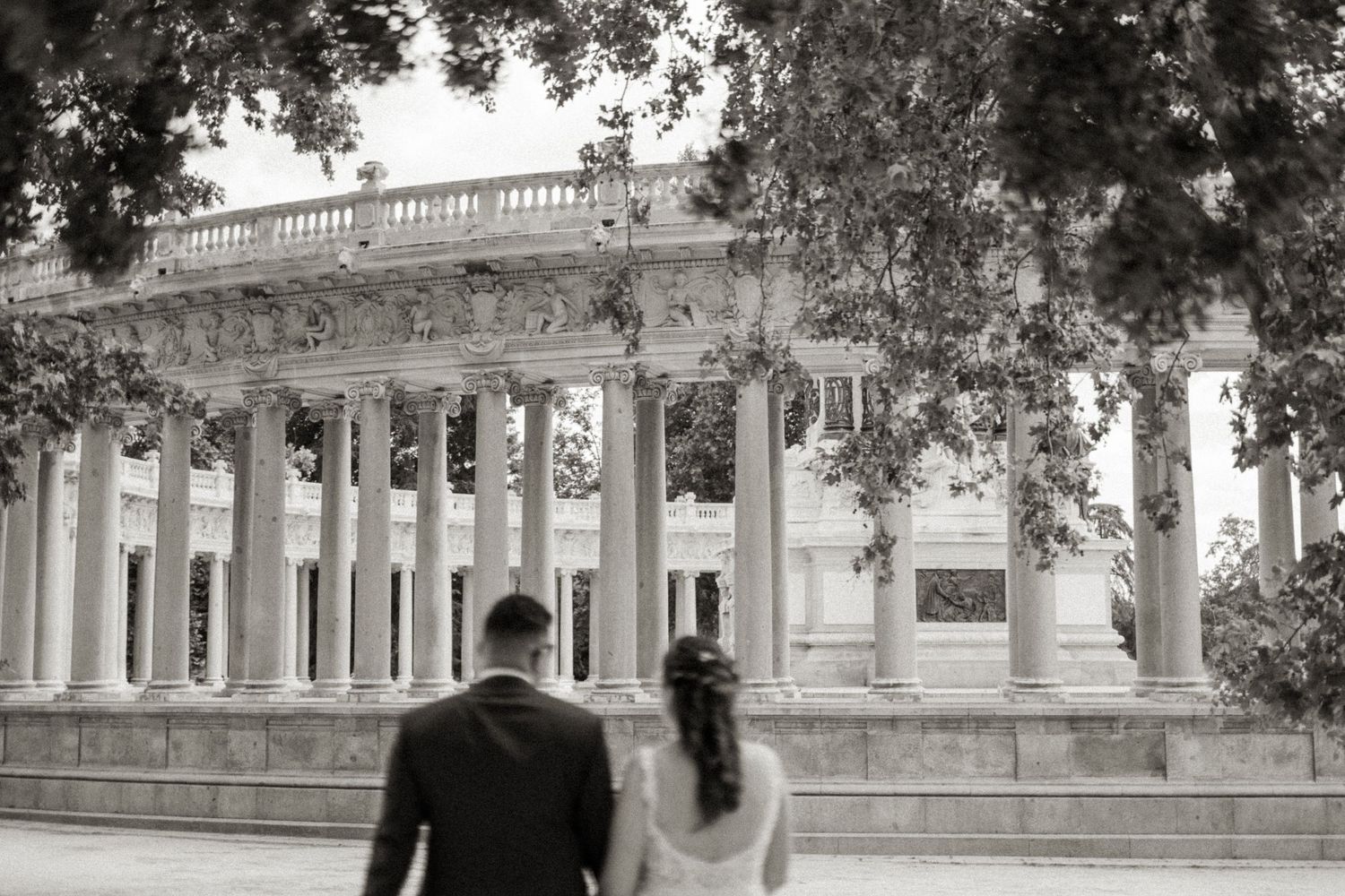 Black and white photo of bride and groom walking toward grand neoclassical building with tall columns.