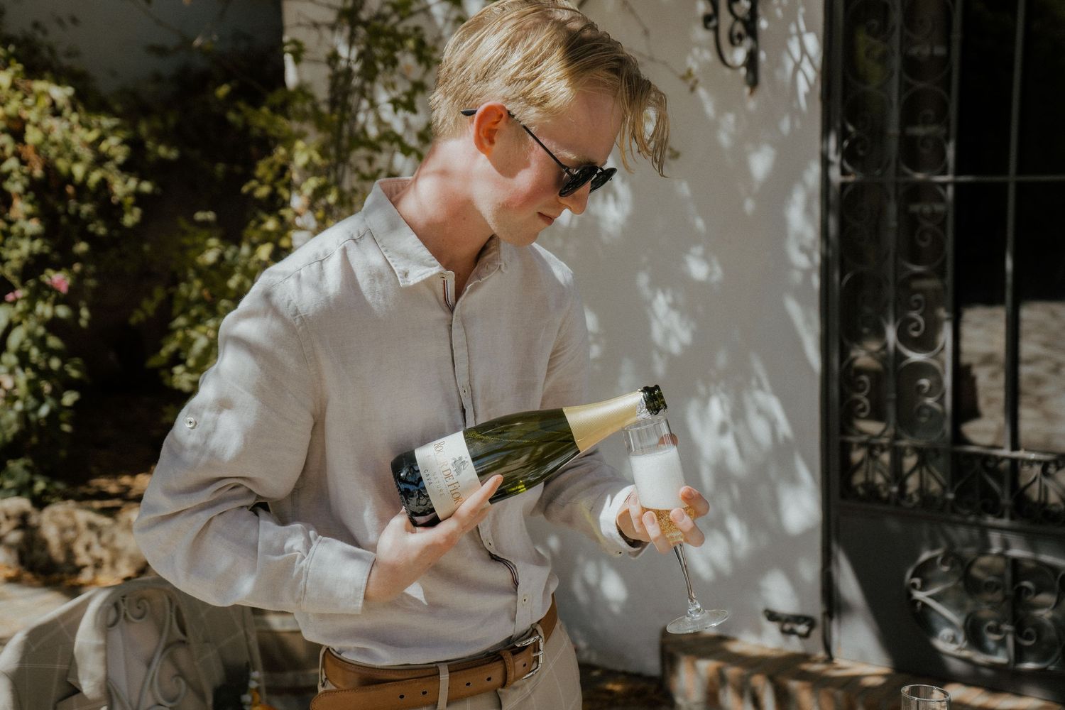 Blonde man in sunglasses opening champagne bottle outdoors in dappled sunlight.