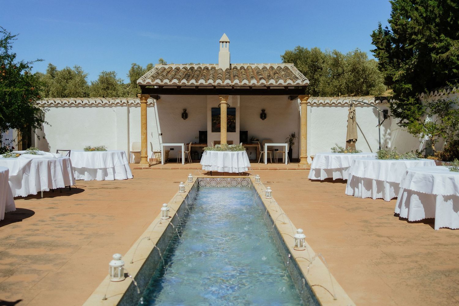 Elegant outdoor wedding venue with long pool, white draped tables, and cypress trees.