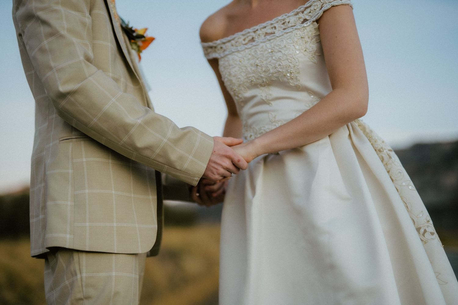 Bride and groom holding hands in wedding attire with golden field and mountains in background.