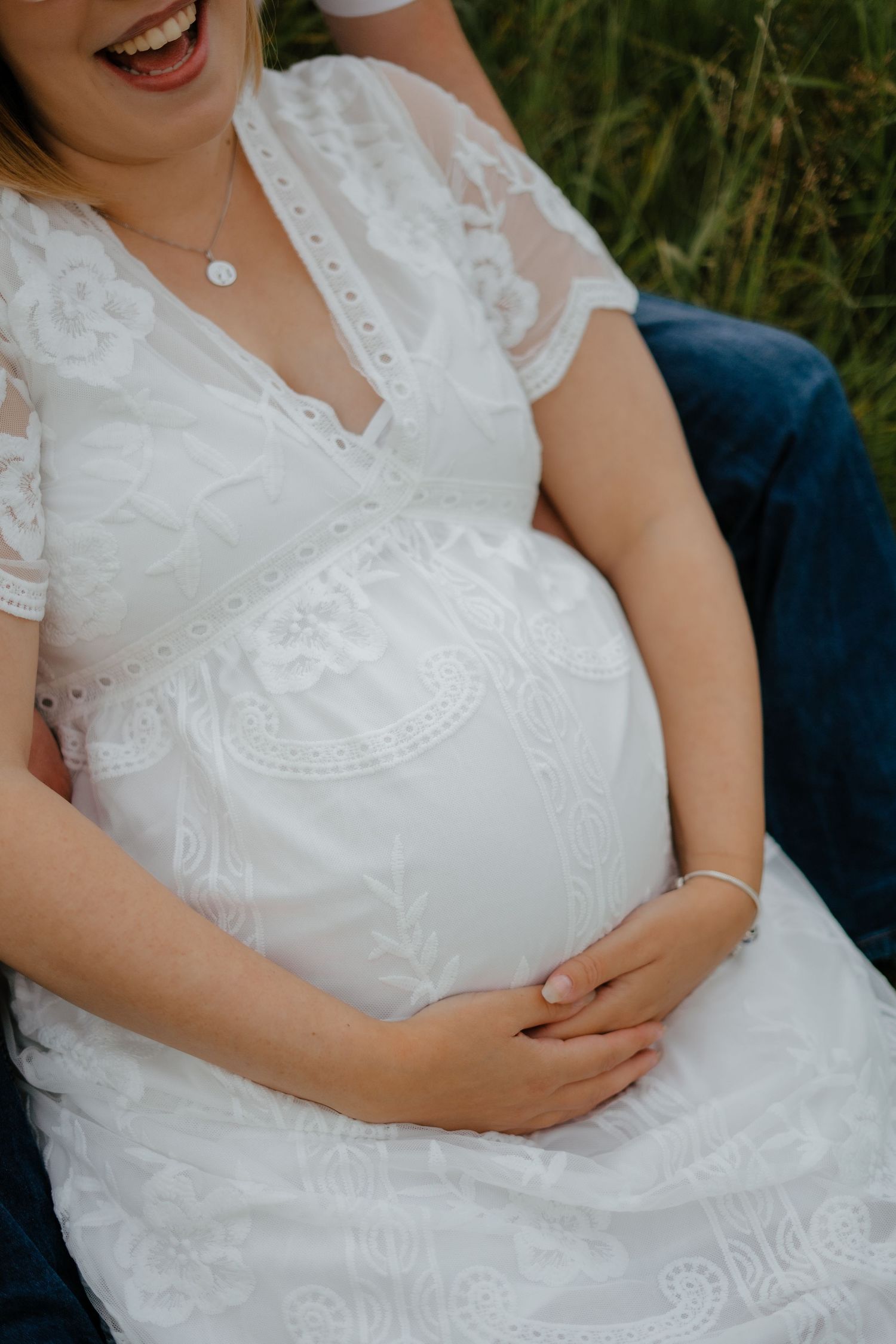 Close-up of a pregnant woman in a white lace dress cradling her baby bump outdoors.