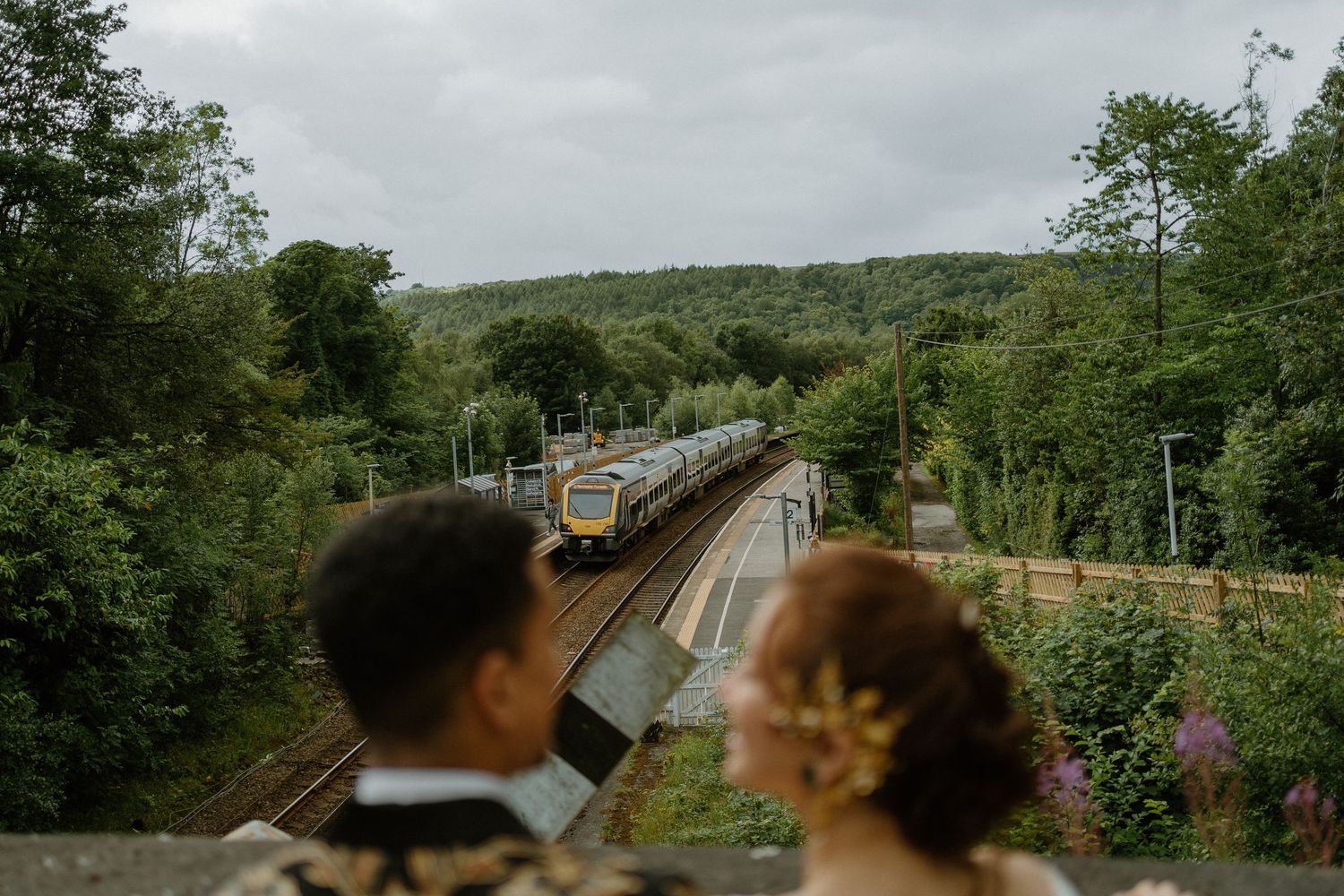 Wedding couple watching a train pass through lush green countryside from a bridge.
