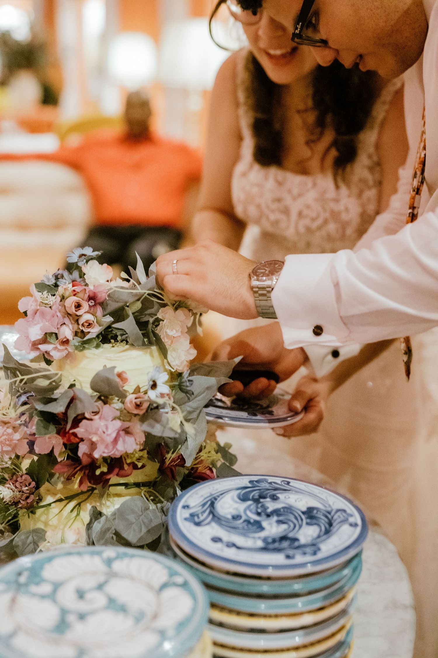 Bride and groom cutting their floral-decorated wedding cake surrounded by colorful ceramic plates.