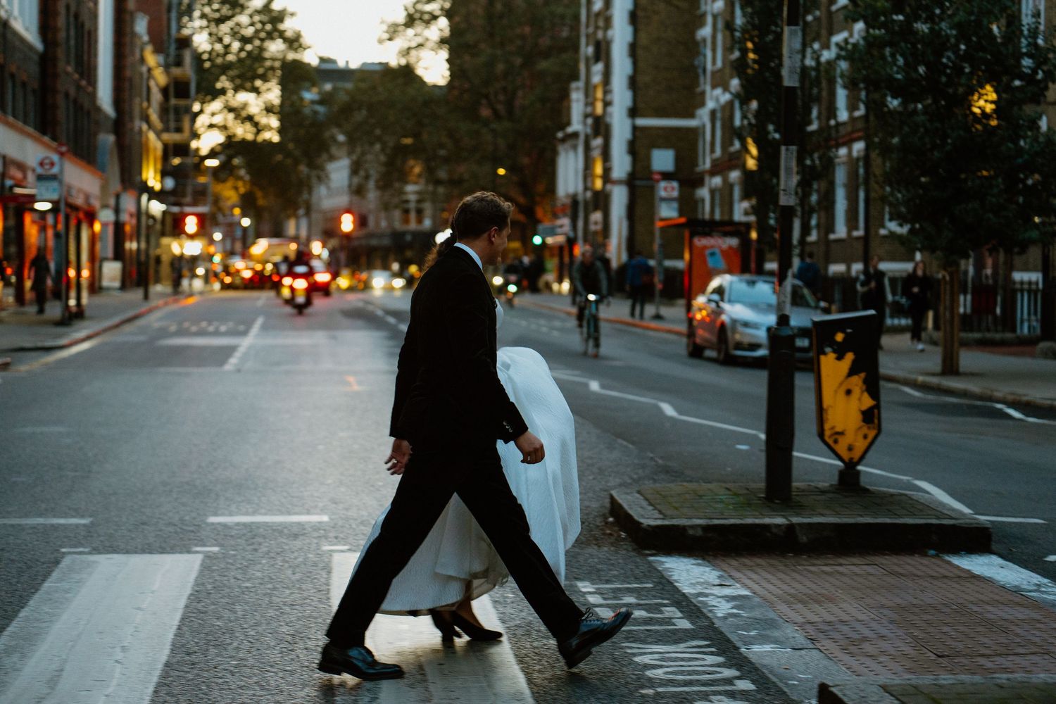 Groom in black suit crossing a busy London street at dusk on his wedding day.