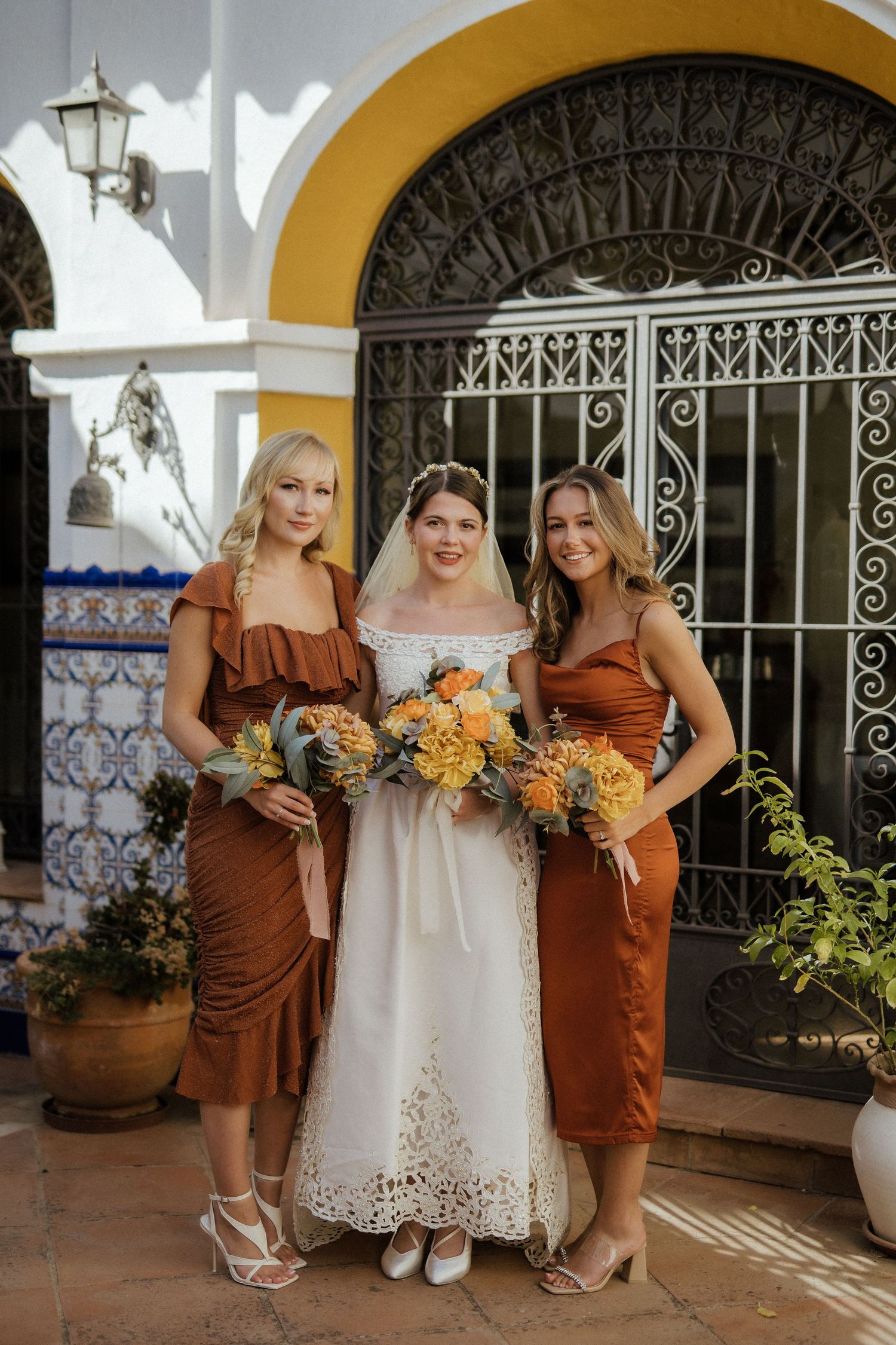 Bride in white lace gown poses with two bridesmaids in rust-colored dresses holding yellow bouquets at a Spanish villa.