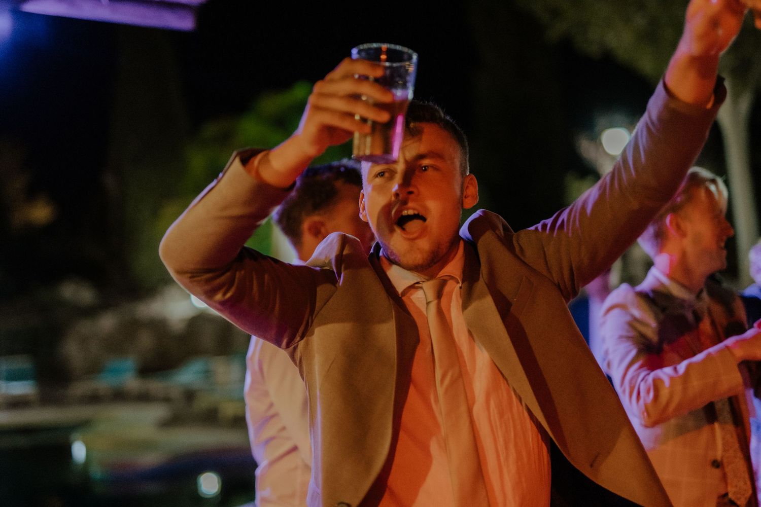Excited wedding guest raising glass in toast at outdoor evening wedding reception.