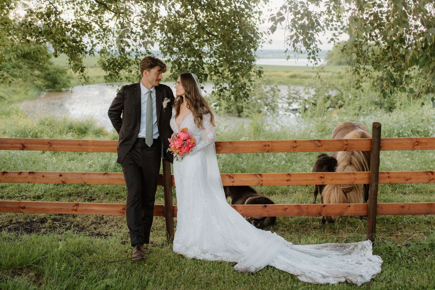 Bride and groom pose by rustic wooden fence with horses in lush green countryside wedding photo.