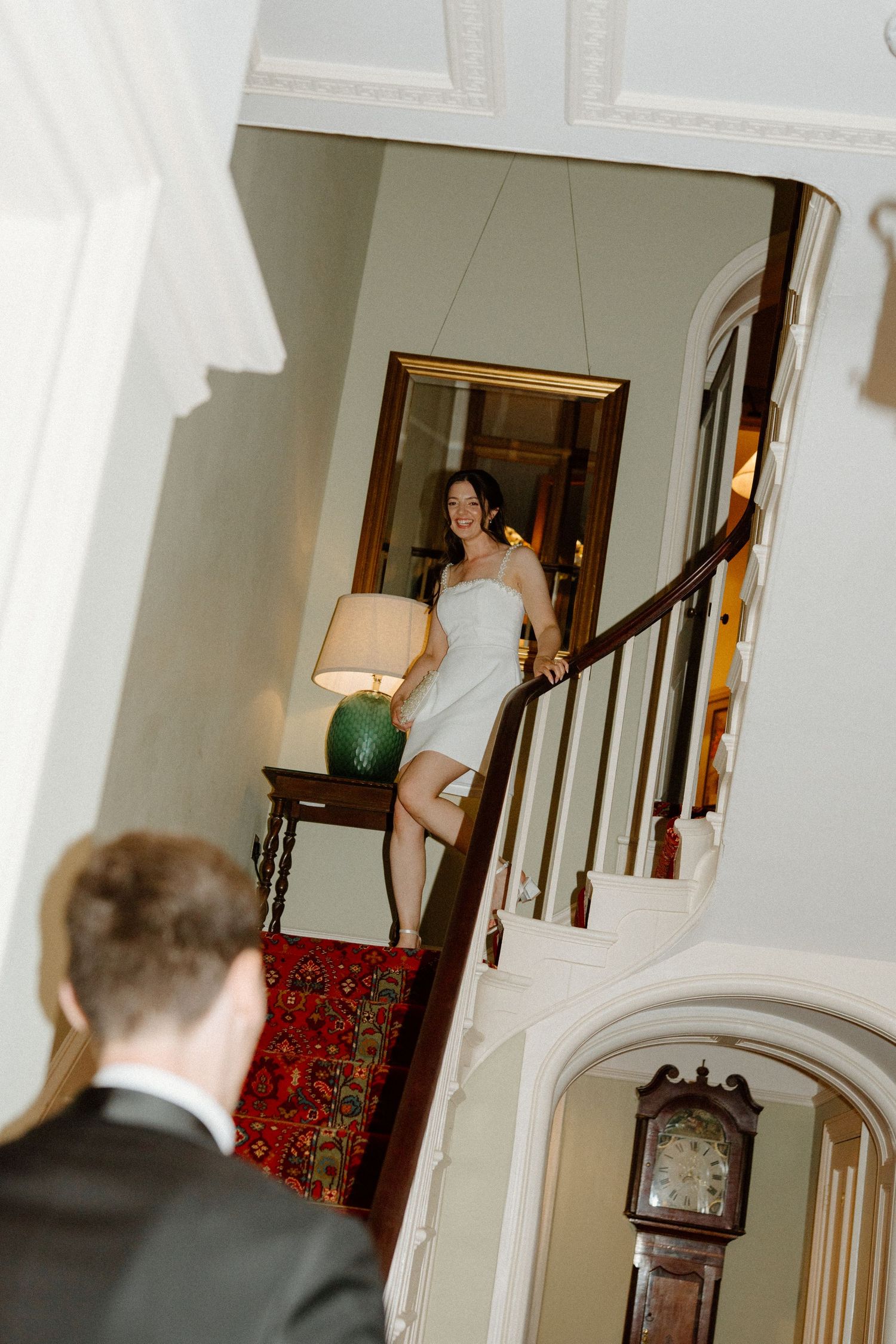 Bride in white dress descending elegant staircase for first look reveal at classic manor house wedding.