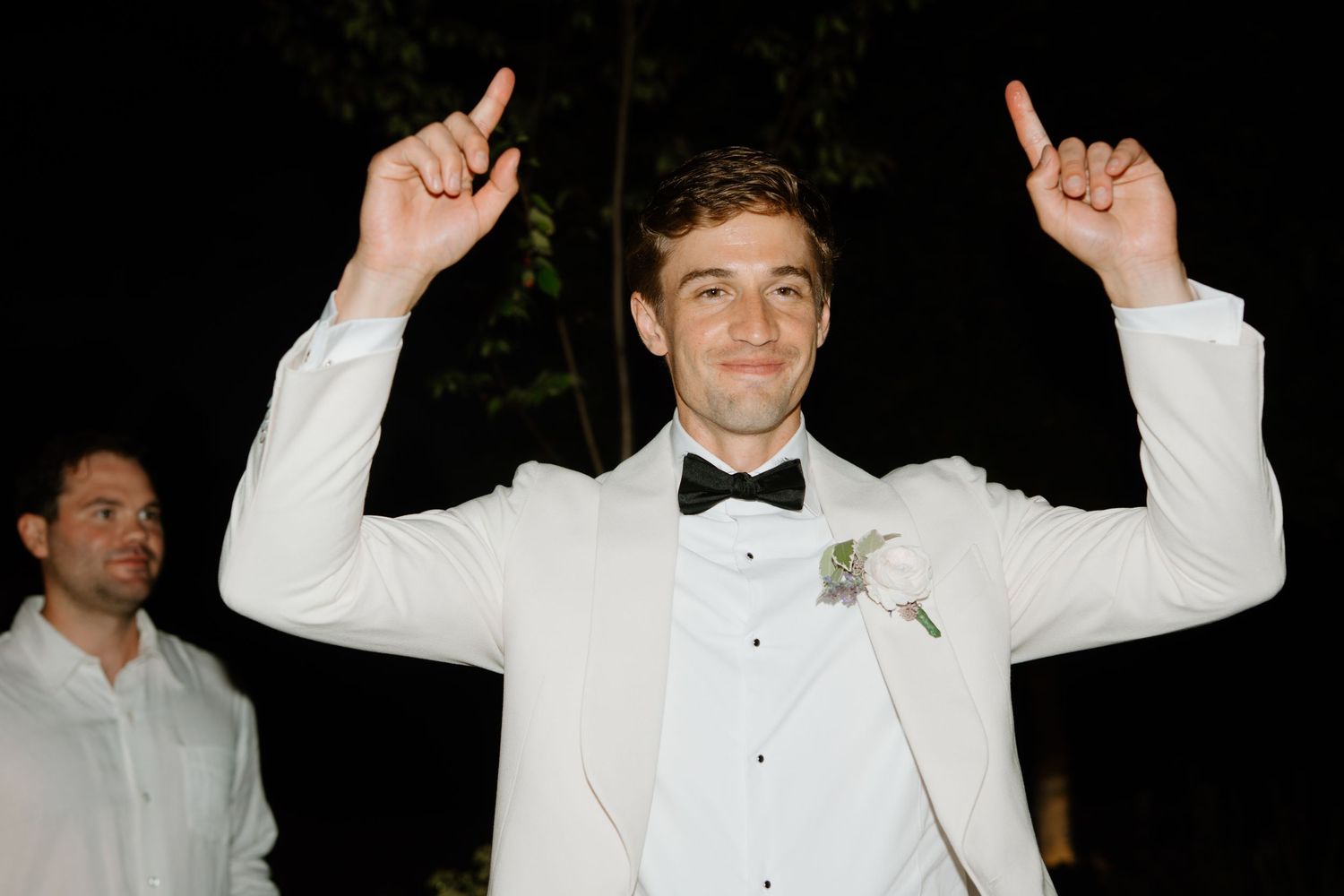 Excited groom in white tuxedo pointing up with both hands at night reception.