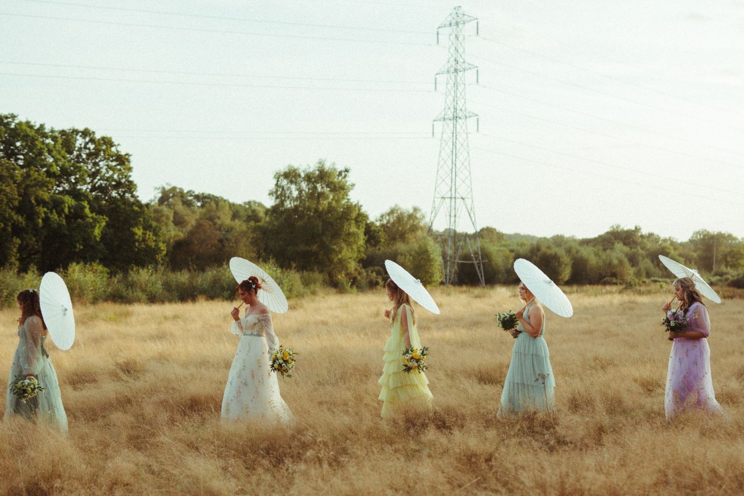 Five bridesmaids in pastel dresses carry white parasols while walking through a golden field.
