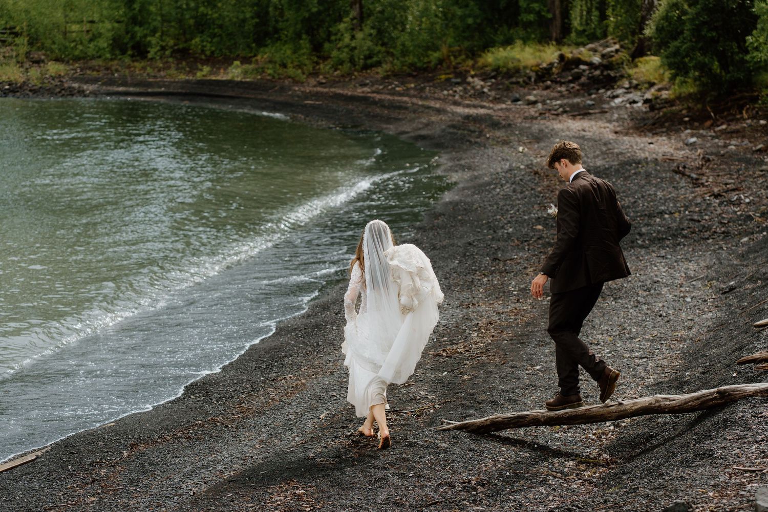 Bride and groom walking barefoot along rocky shoreline with veil flowing in the breeze.