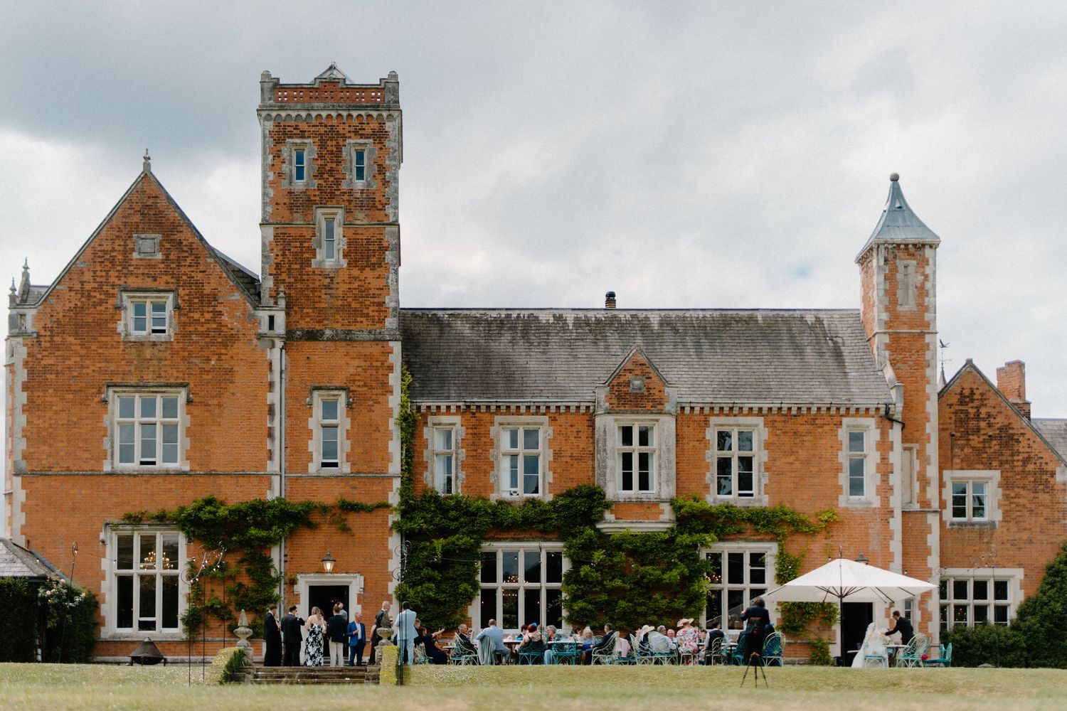 Grand red brick English manor house with gothic architecture surrounded by wedding guests on the lawn.
