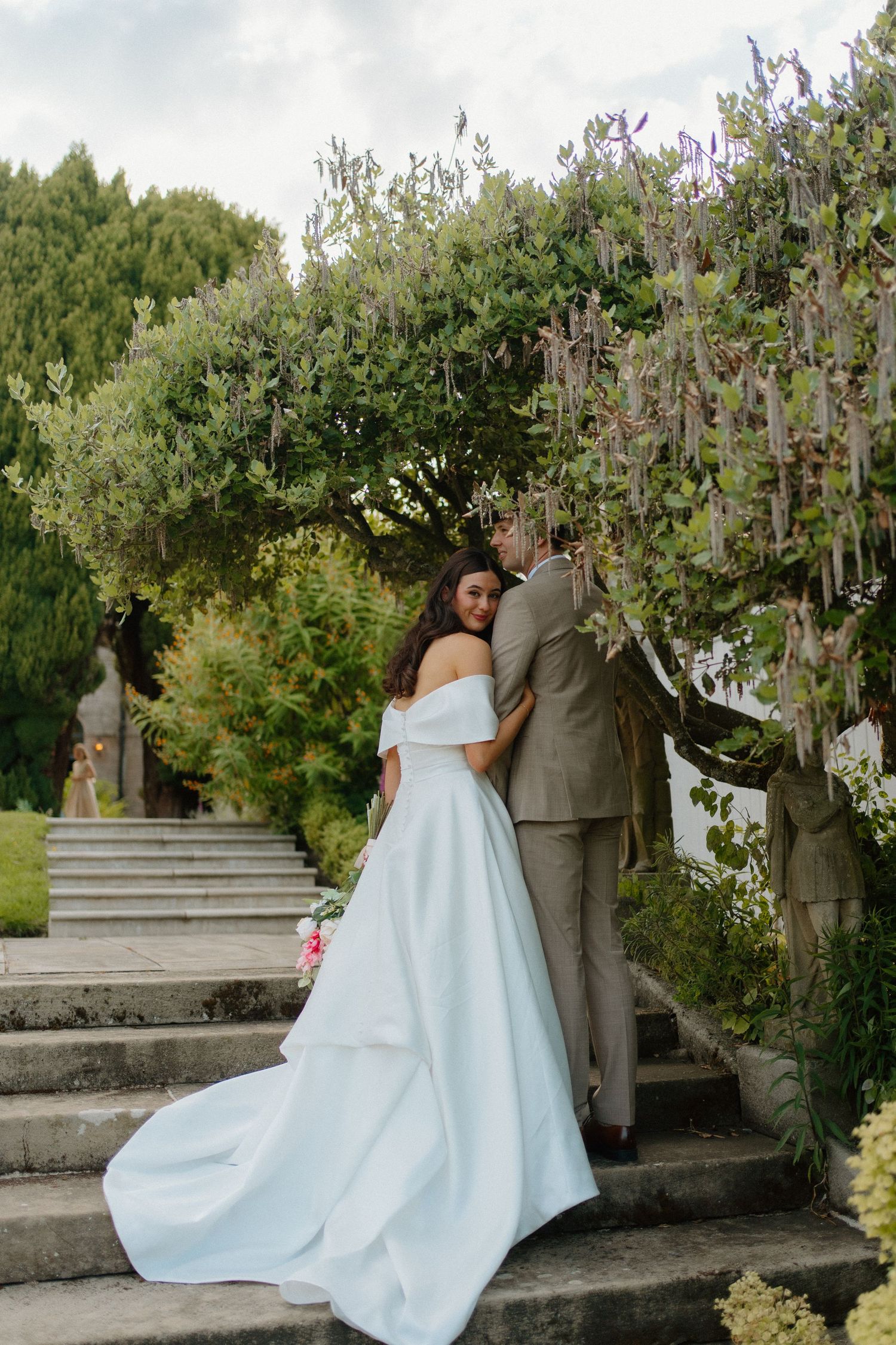 Bride in a flowing white off-shoulder gown and groom in tan suit posing on stone steps beneath a wisteria archway.
