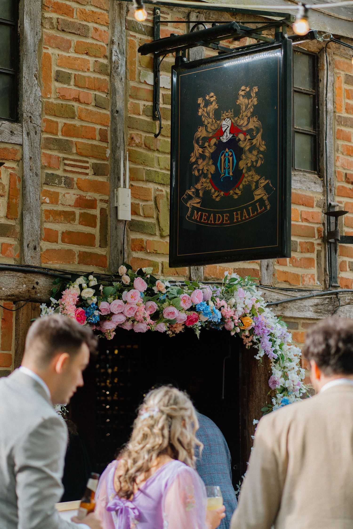 Wedding guests gather beneath a floral arch and ornate pub sign at historic Meade Hall with exposed brick walls.