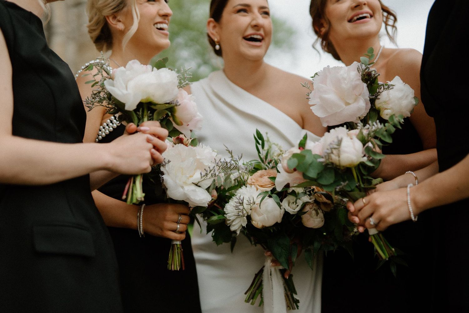 Bridesmaids hold white and peach floral bouquets while sharing a candid laughing moment at a wedding celebration.
