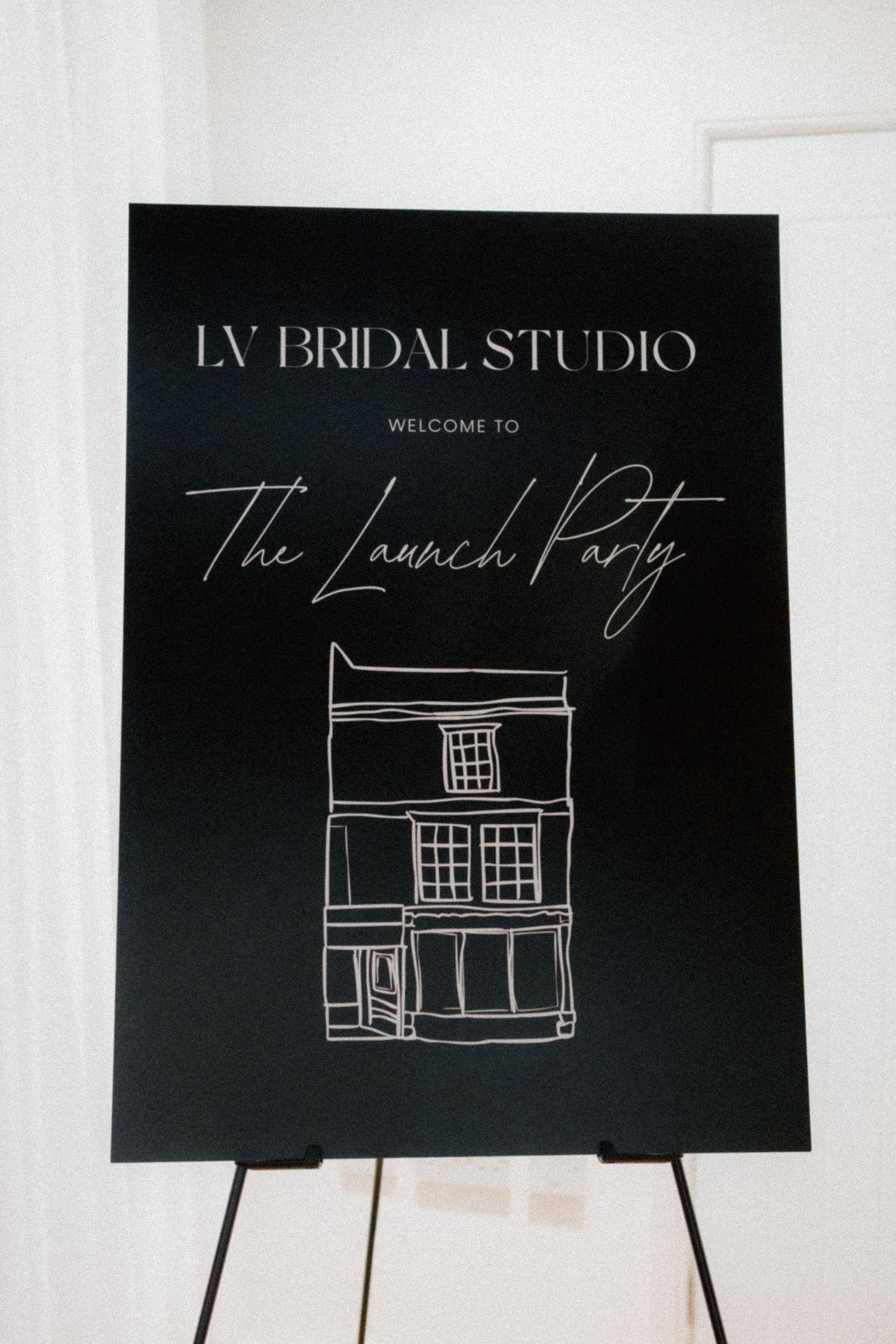 A black welcome sign for LV Bridal Studio with elegant script text and a simple sketched building design.