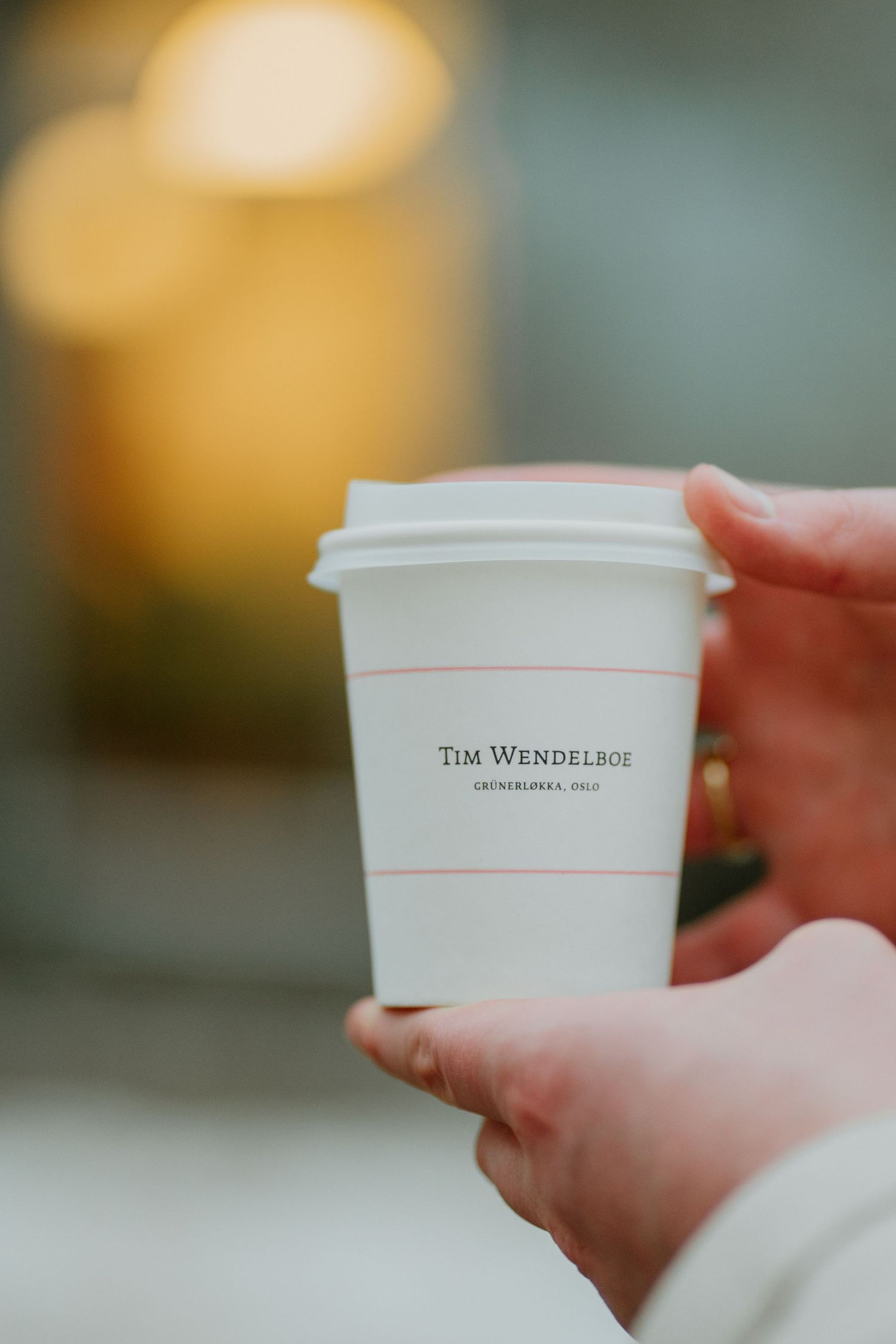Someone holding a white Tim Wendelbo takeaway coffee cup with blurred warm background lighting.