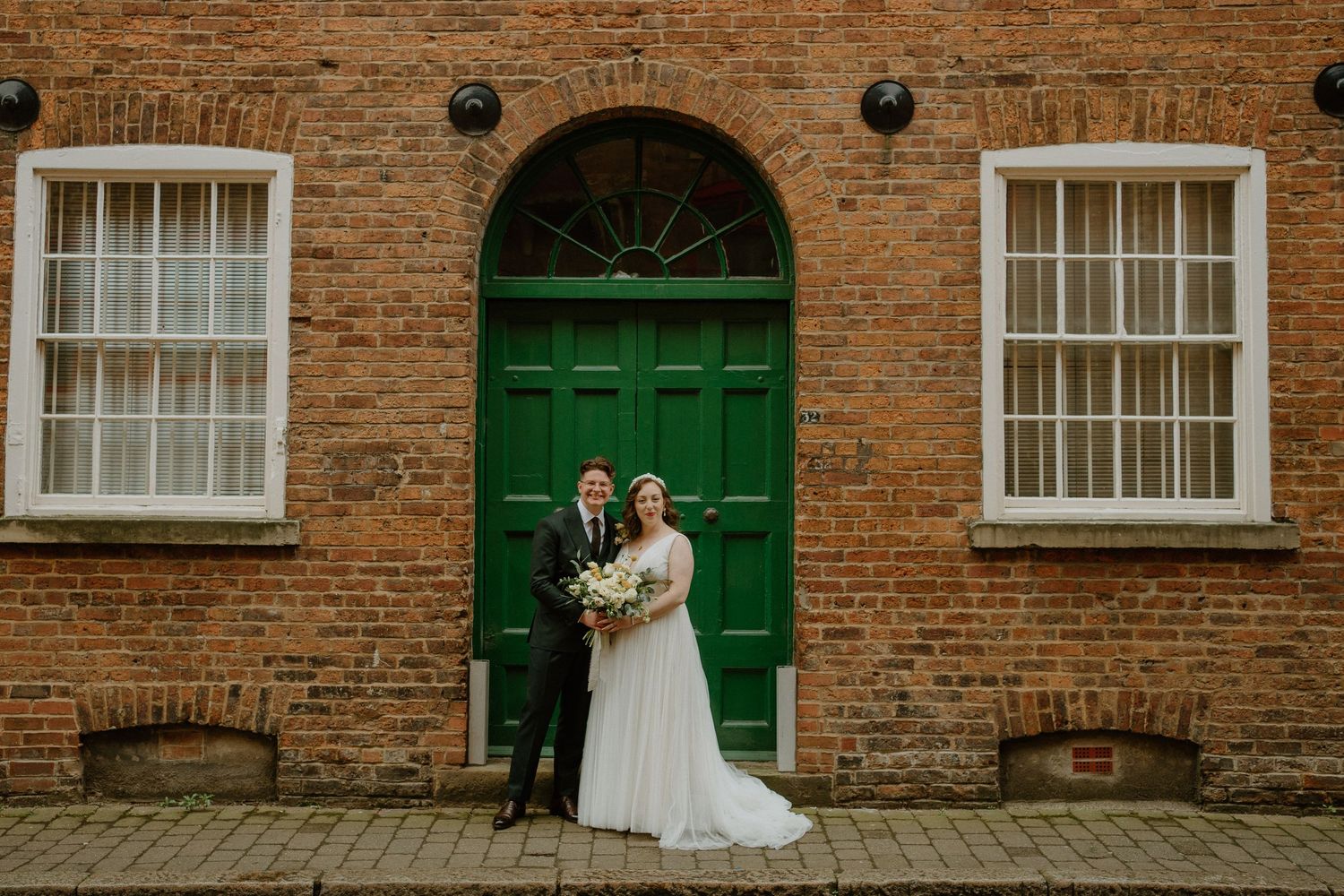 Bride and groom posing in front of a green arched door on a brick building on their wedding day.