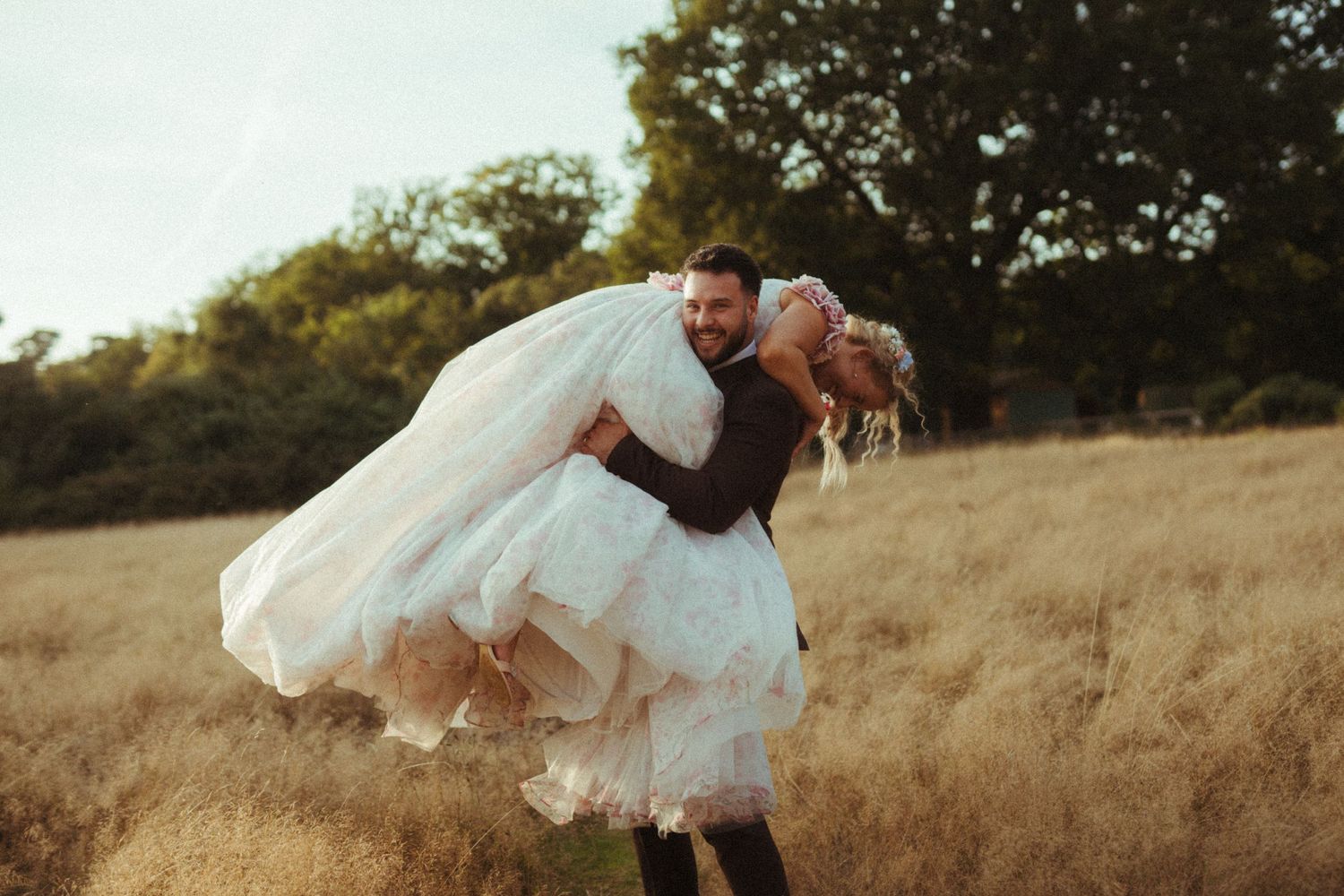 A couple in wedding attire share a romantic dip pose in a golden field at sunset.