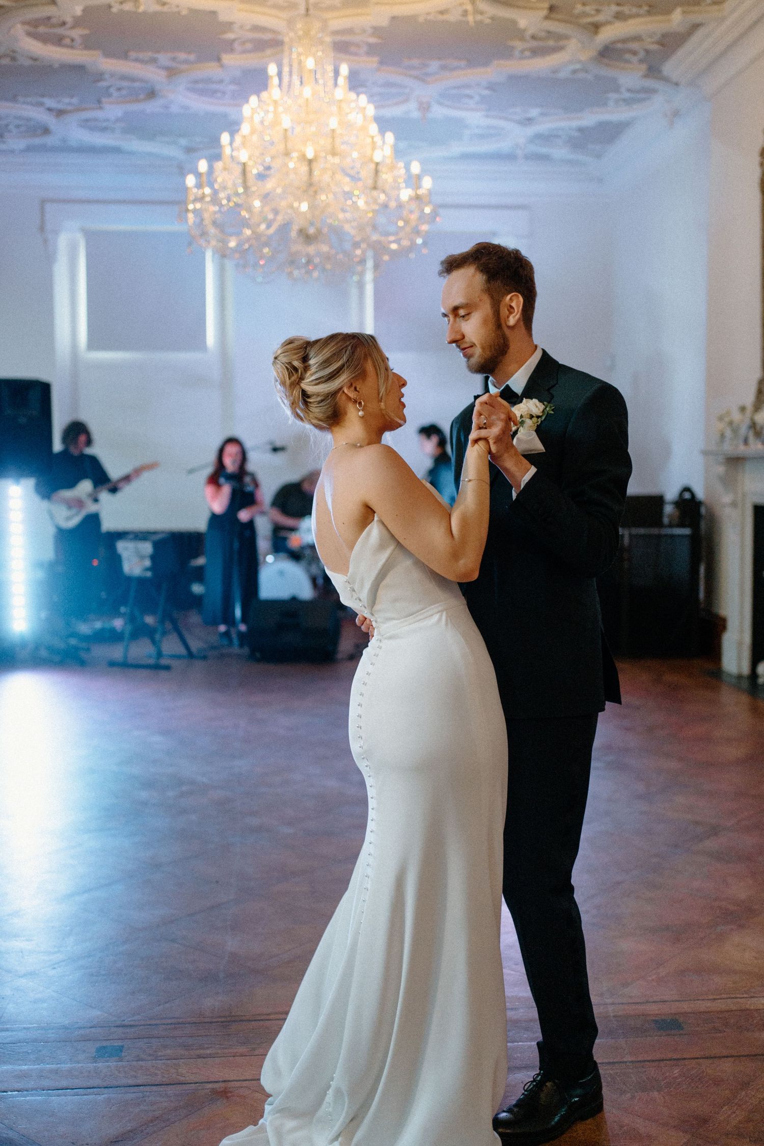Bride and groom share romantic first dance under crystal chandelier with live band performing in background.