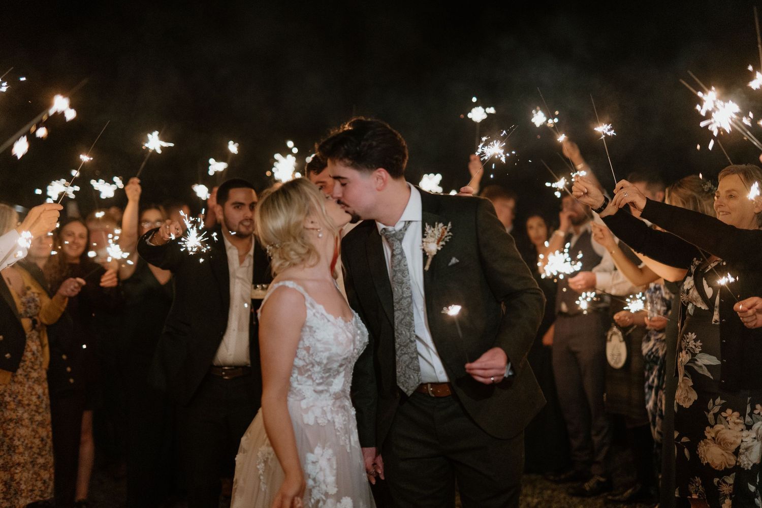 Bride and groom sharing a romantic kiss surrounded by guests holding sparklers during nighttime wedding exit.