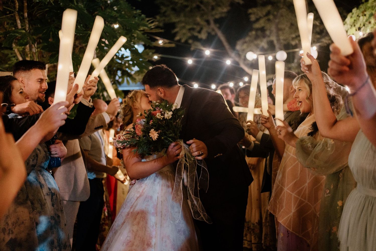 Bride and groom sharing a kiss while guests wave glowsticks during a festive wedding sendoff.