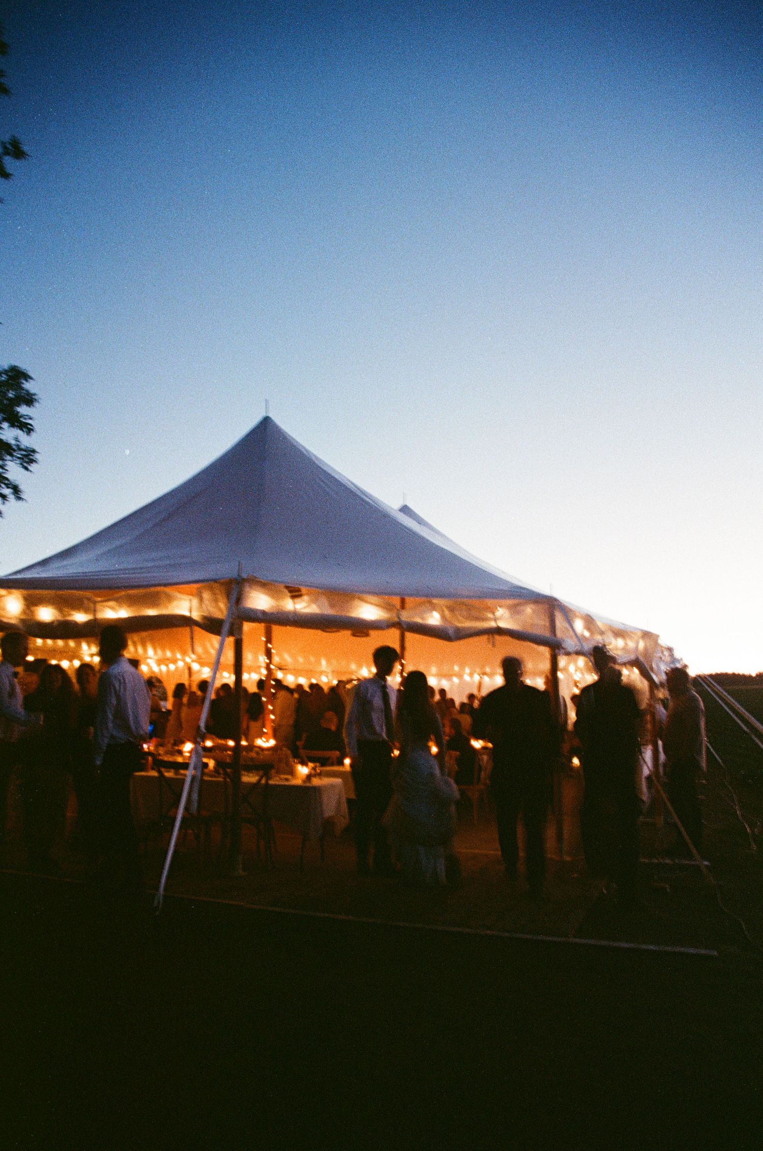 Glowing white wedding tent at dusk with string lights and guests mingling under a clear blue sky.