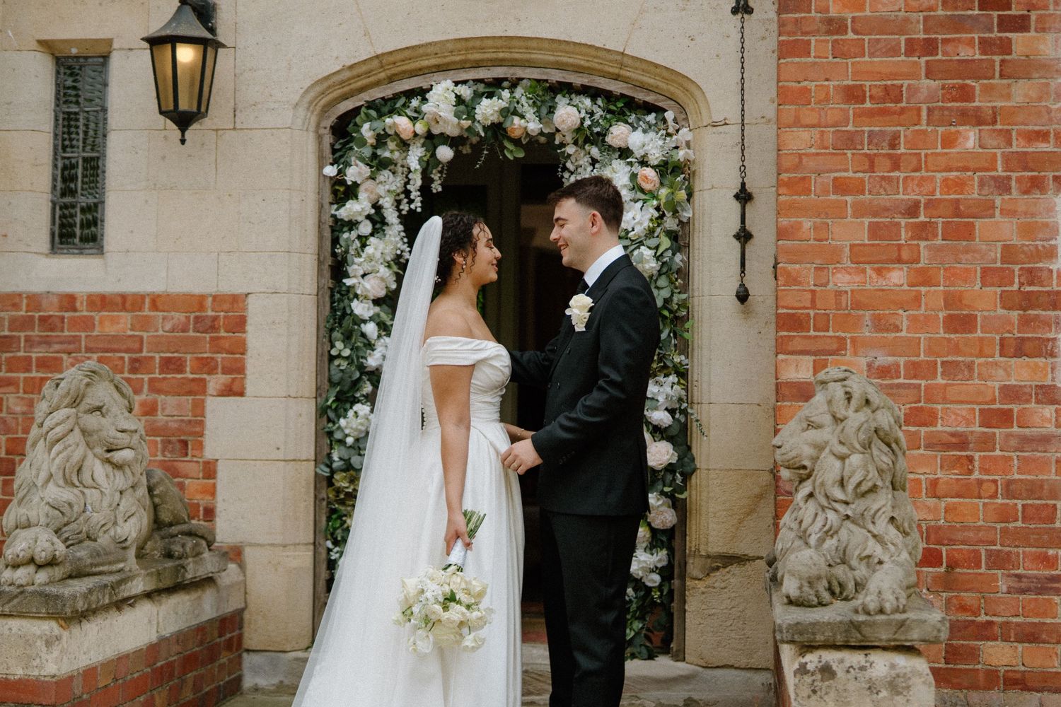 Bride and groom holding hands before floral arch at brick venue.