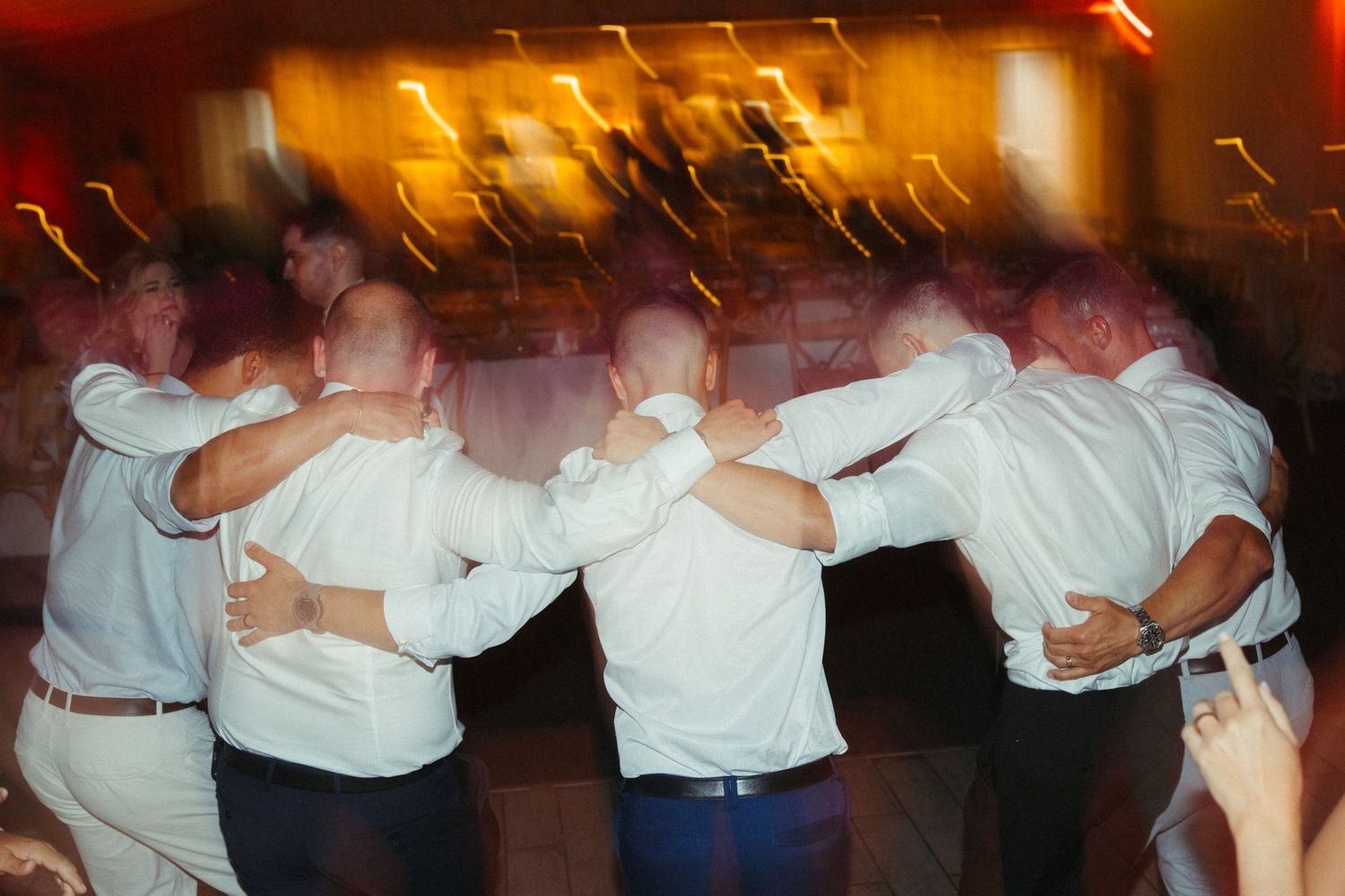 Group of men in white shirts dance together in circle on wedding reception dance floor.